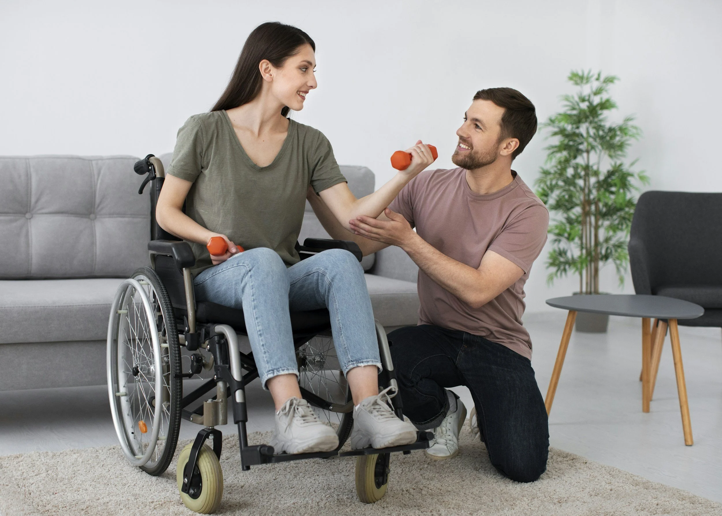 A woman in a wheelchair lifting a small orange dumbbell while a man kneels beside her holding her arm, smiling.