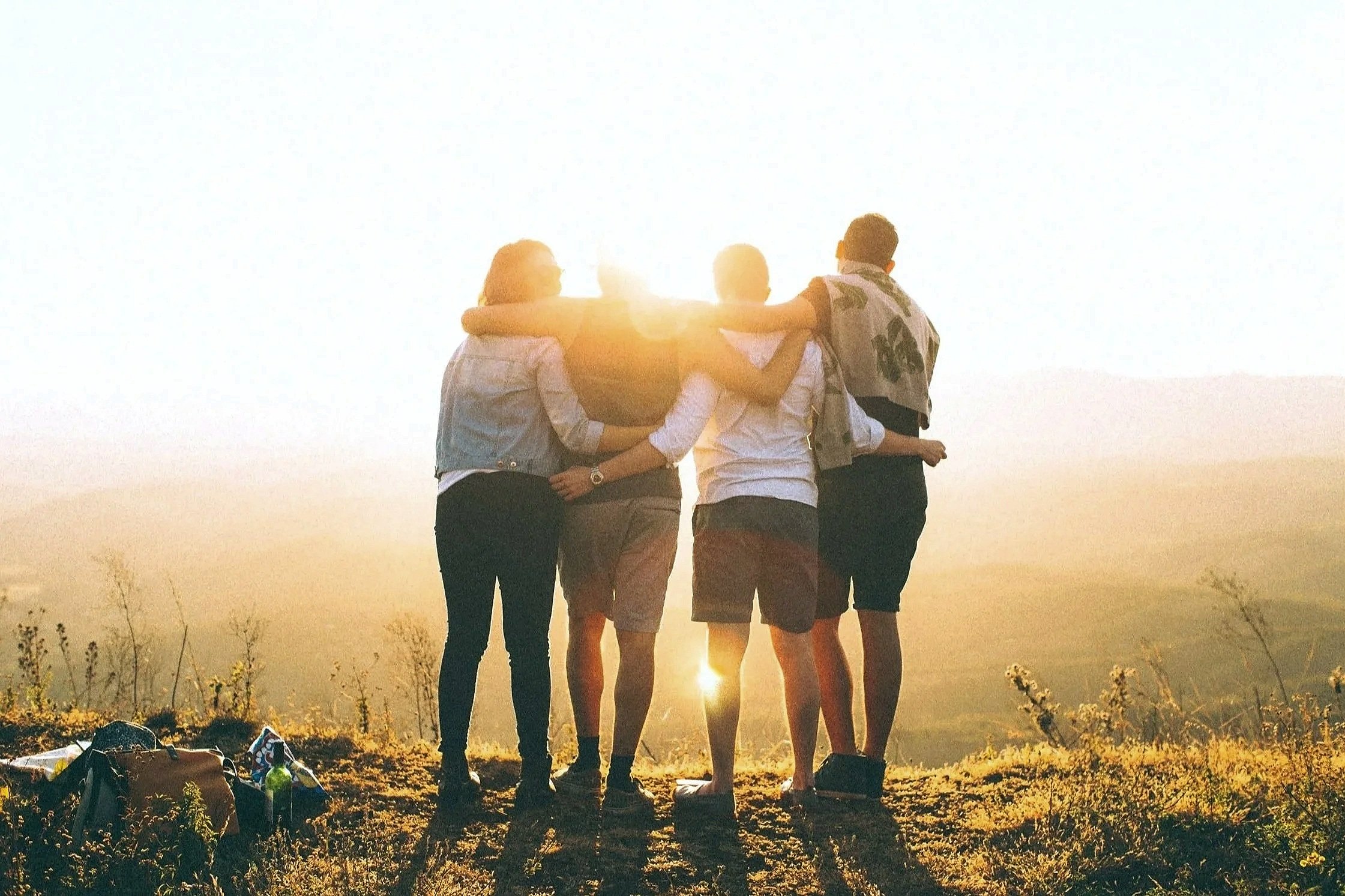 Group of five friends standing together outdoors during sunset, with arms around each other, overlooking a scenic landscape.