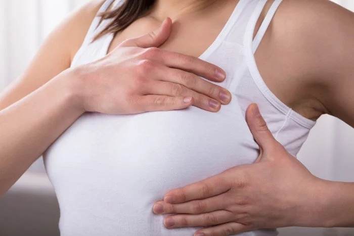 A woman wearing a white tank top holding her chest and stomach, possibly experiencing discomfort.