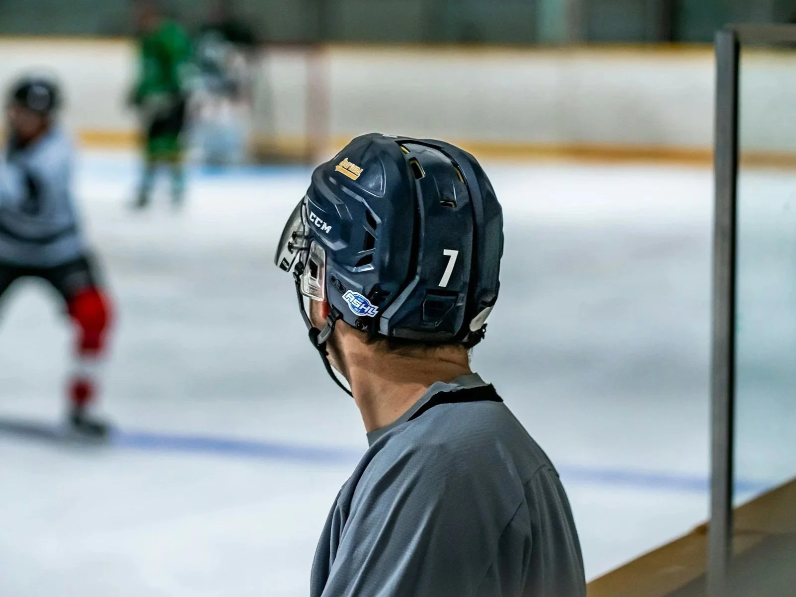 A hockey coach wearing a helmet and gray shirt standing near the rink, watching players on the ice during a game.