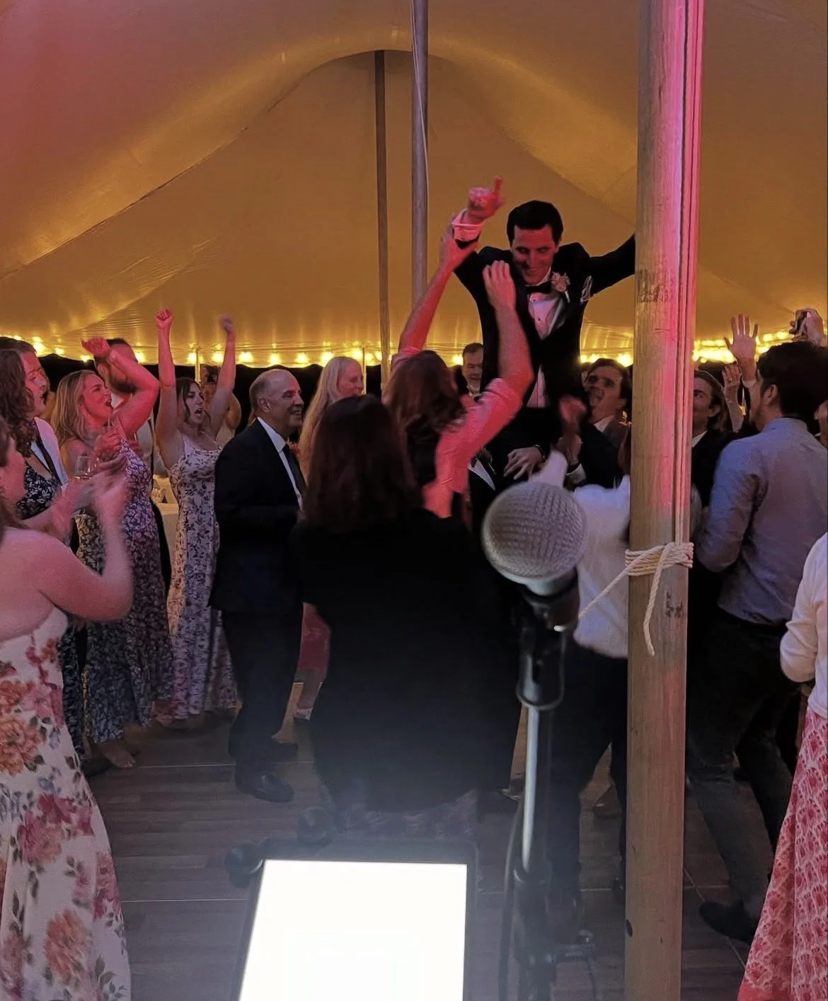People dancing and celebrating under a large yellow tent, with one person lifted by another, at a festive event.