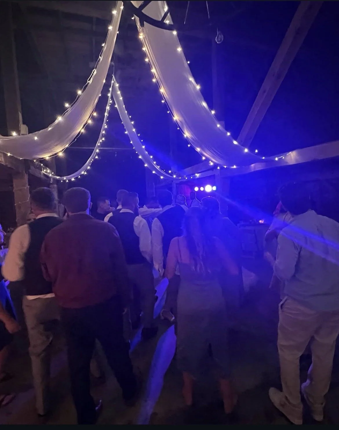 People dancing and socializing at an indoor party with string lights and draped fabric decorations hanging from the ceiling.
