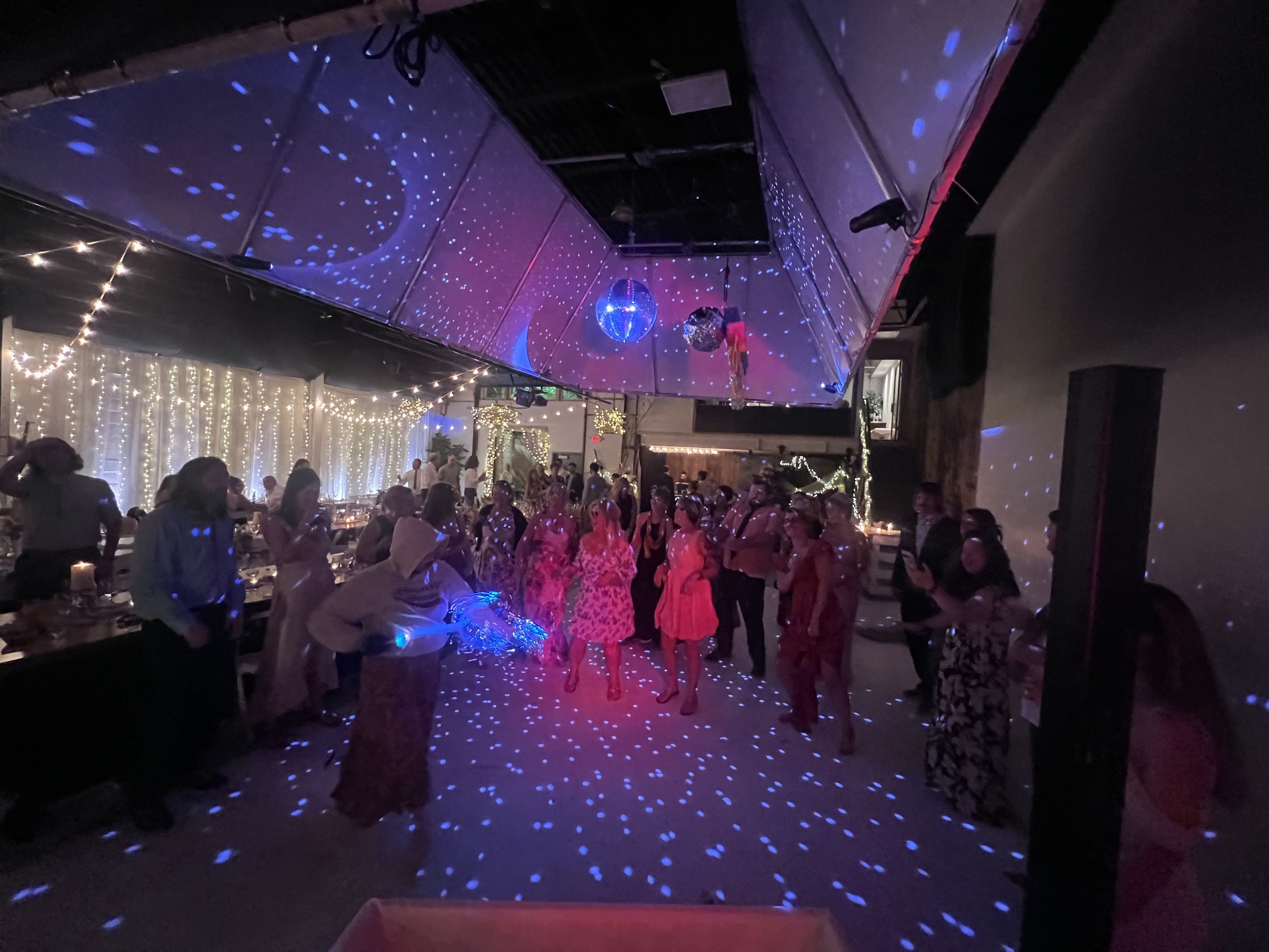 People dancing and socializing at a party with colorful lighting, fairy lights, and disco balls.