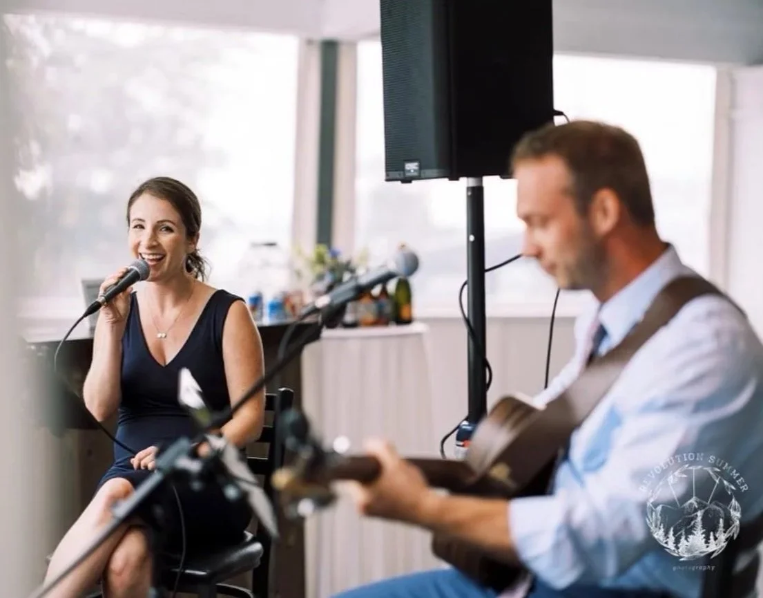 A woman singing into a microphone and a man playing guitar in a bright, casual indoor setting.