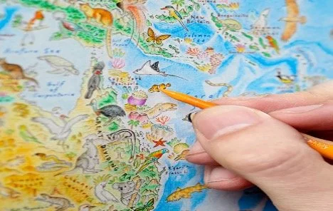 Close-up of a hand pointing with an orange pencil at a colorful illustrated map by artist Anton Thomas.