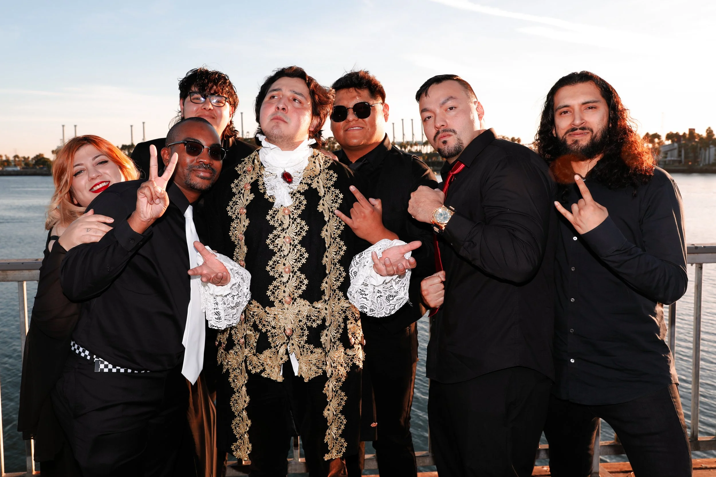 Group of seven people dressed in black and ornate costumes posing outdoors near water at sunset.