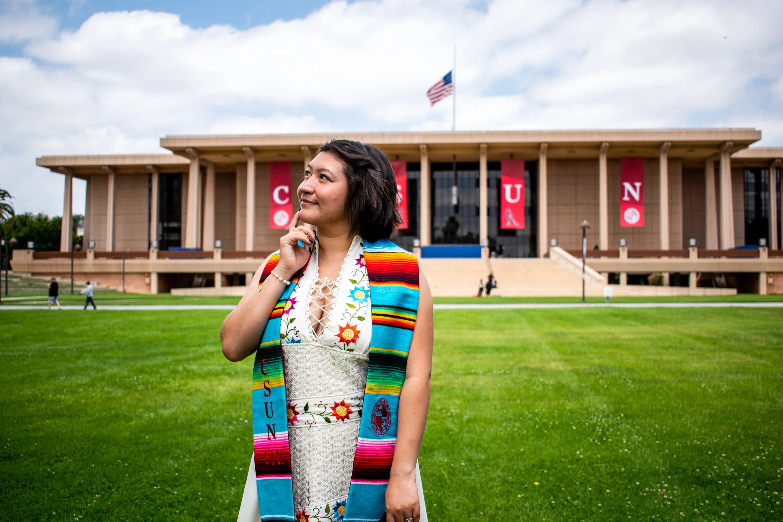 A woman in traditional Mexican attire standing in front of a university building with American flags, wearing a colorful scarf and dress, with a thoughtful expression.