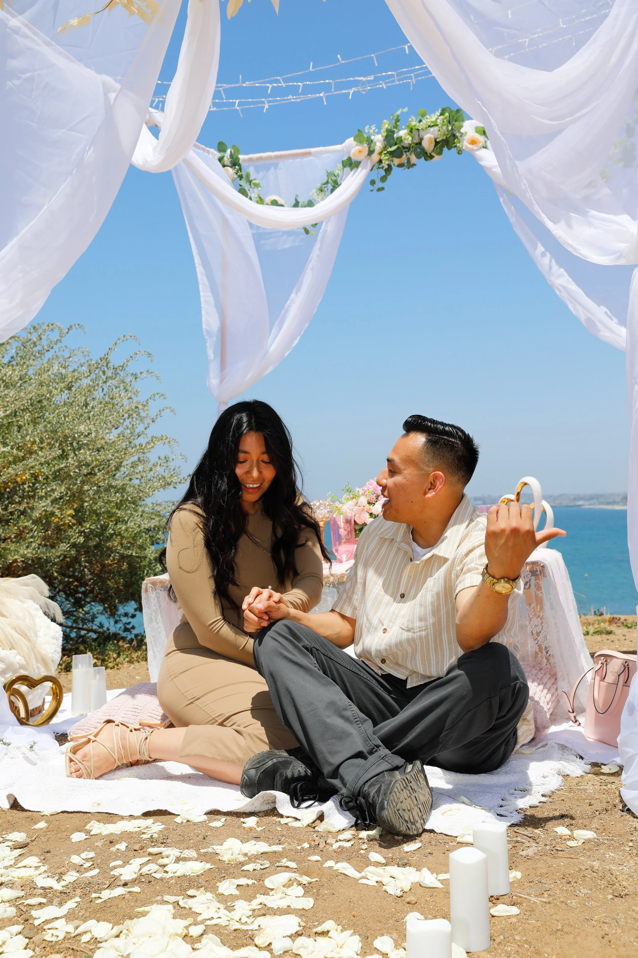 A couple sitting on a blanket during an outdoor wedding ceremony by the ocean. The scene features white drapery, floral decorations, candles, and a blue sky.