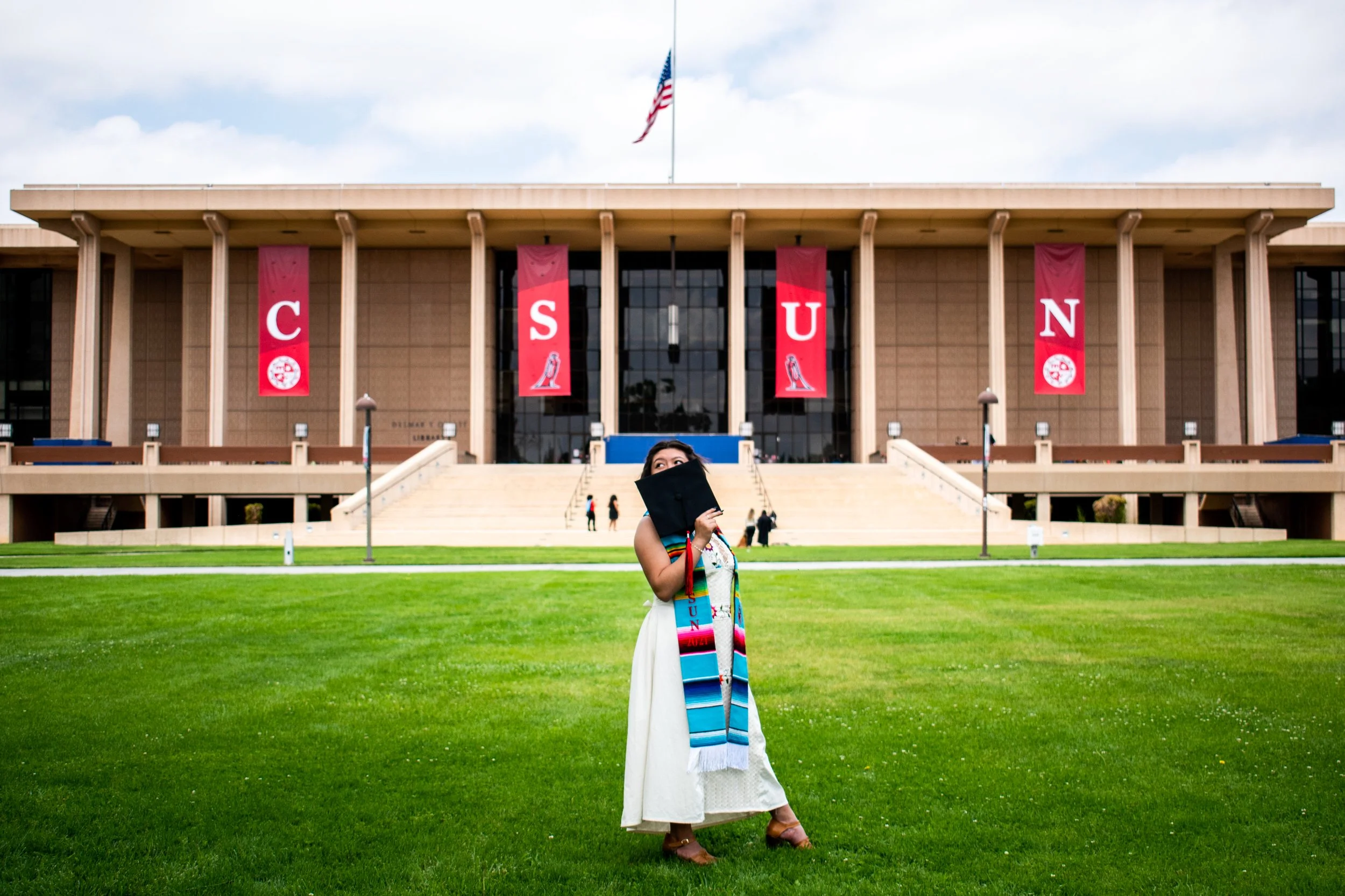 A woman in a white dress and graduation stole standing on a green lawn, holding a black diploma cover in front of her face, in front of a large university building with red banners that spell 'CSUN' and an American flag on top.