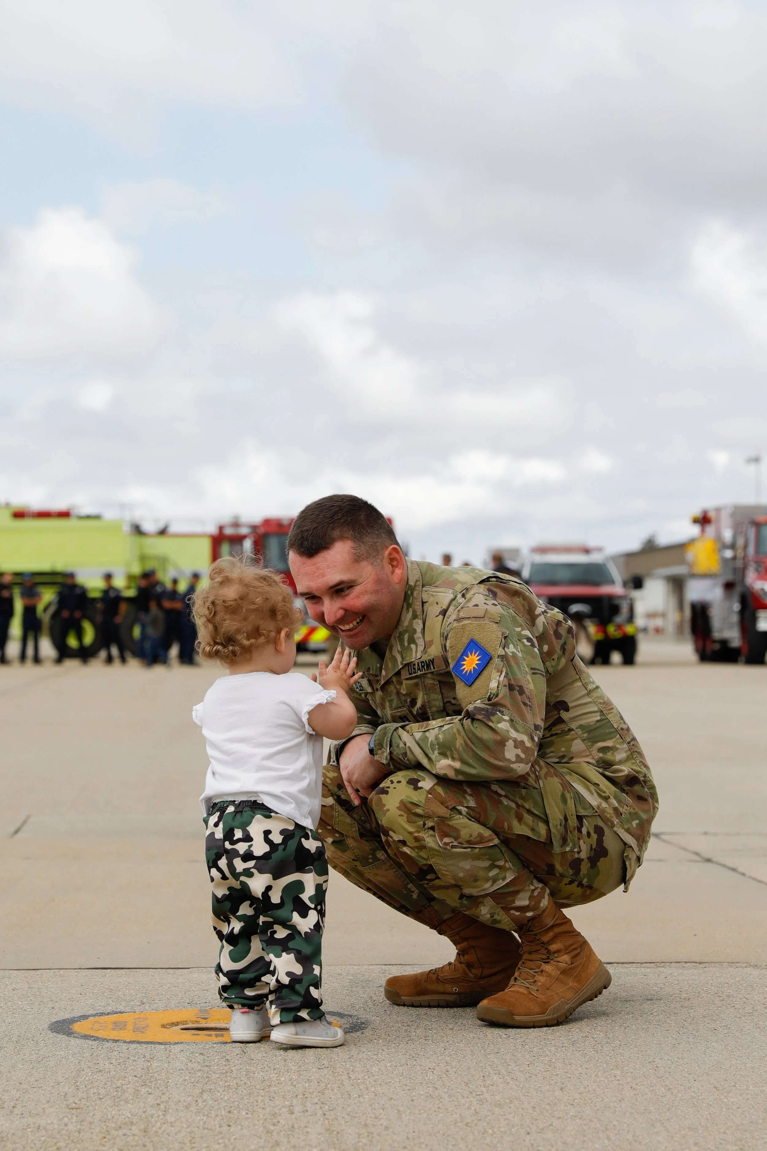 A soldier in camouflage uniform kneels and smiles at a young girl with curly hair, who is touching his face. The background shows emergency vehicles and personnel, suggesting a public event or airbase.