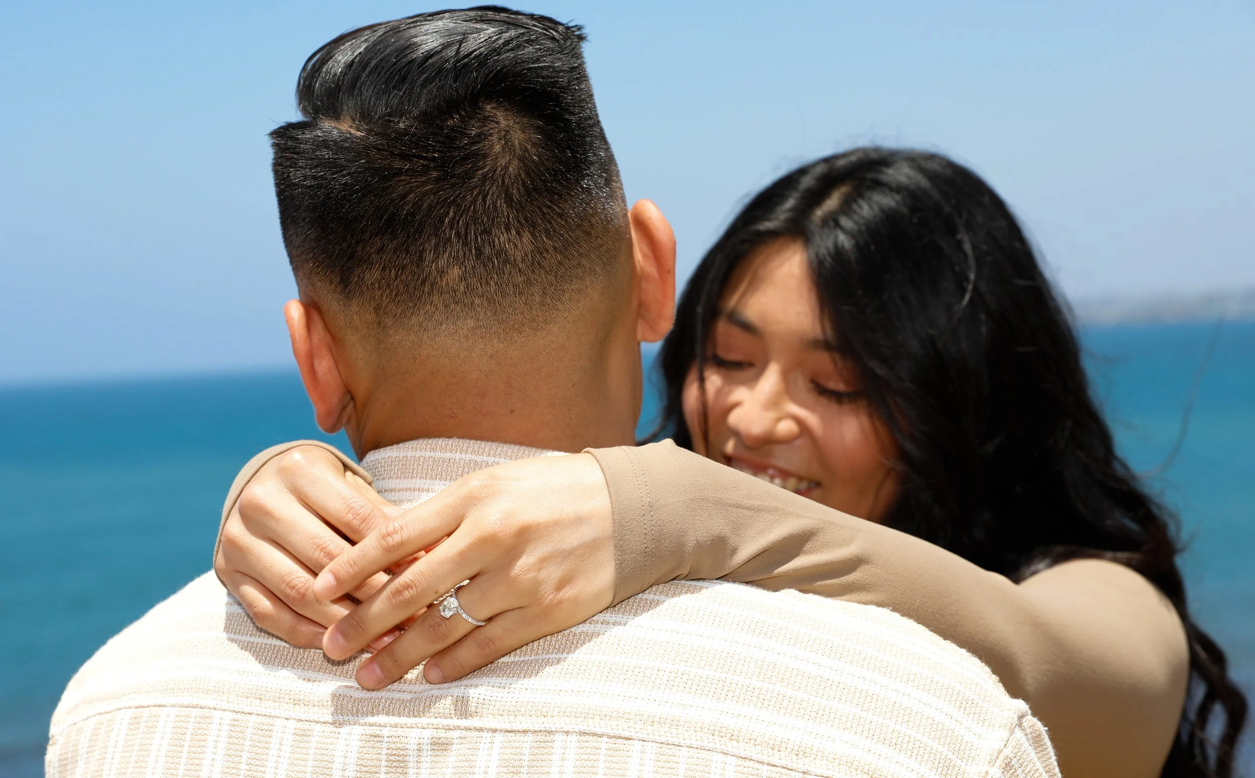 A couple hugging, with a beach and ocean in the background. The woman is smiling, showing her engagement ring.