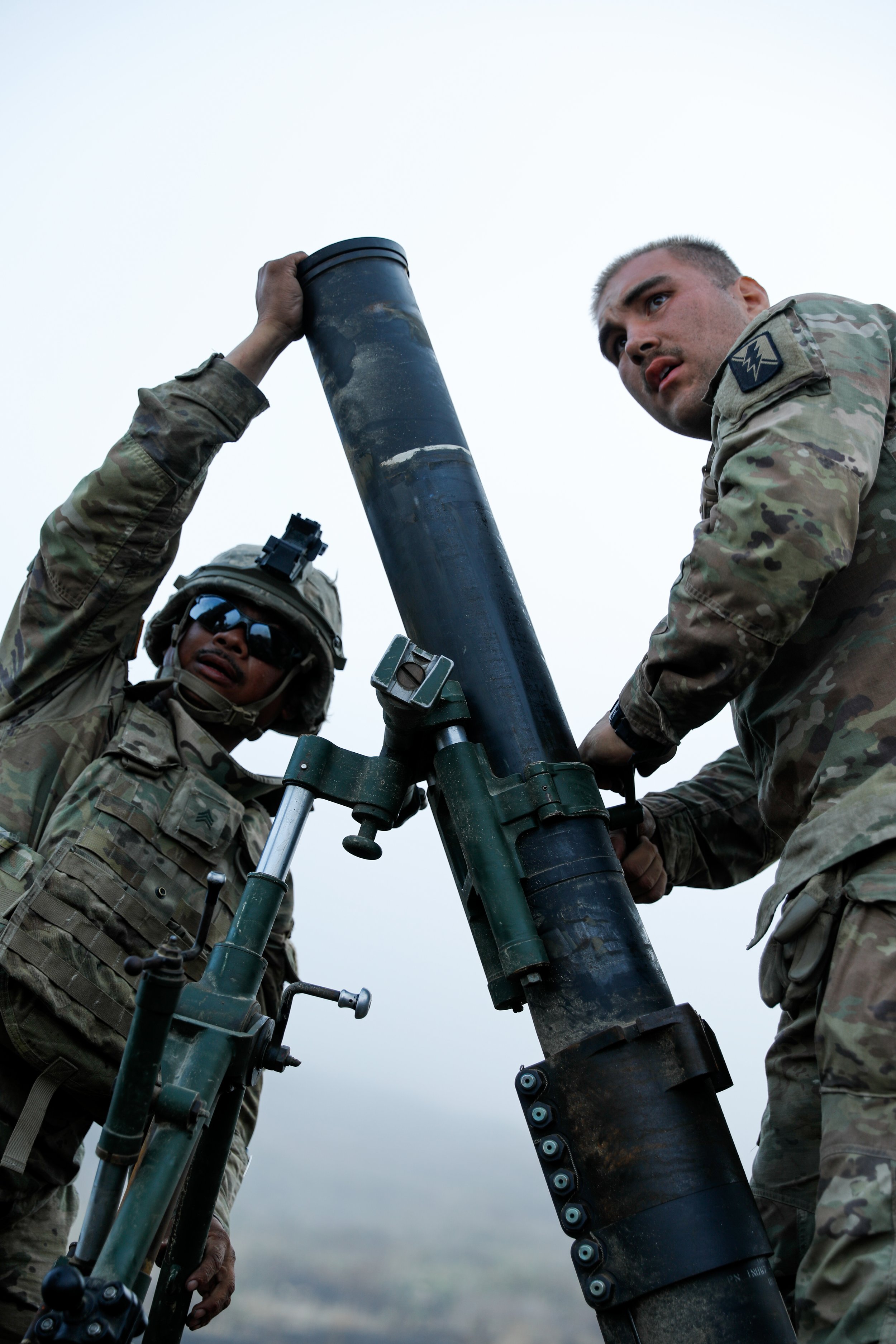 Two soldiers in camouflage uniforms and helmets are adjusting a large artillery shell on a military weapon outdoors on a cloudy day.