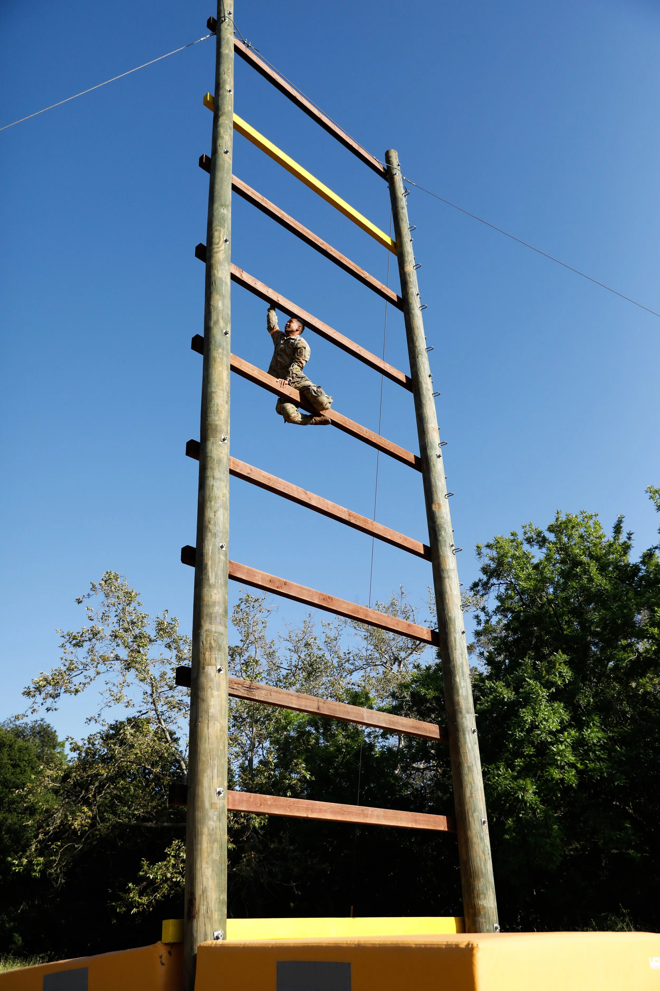 A person in military uniform climbing an outdoor wooden and metal obstacle course ladder, with trees and a clear blue sky in the background.