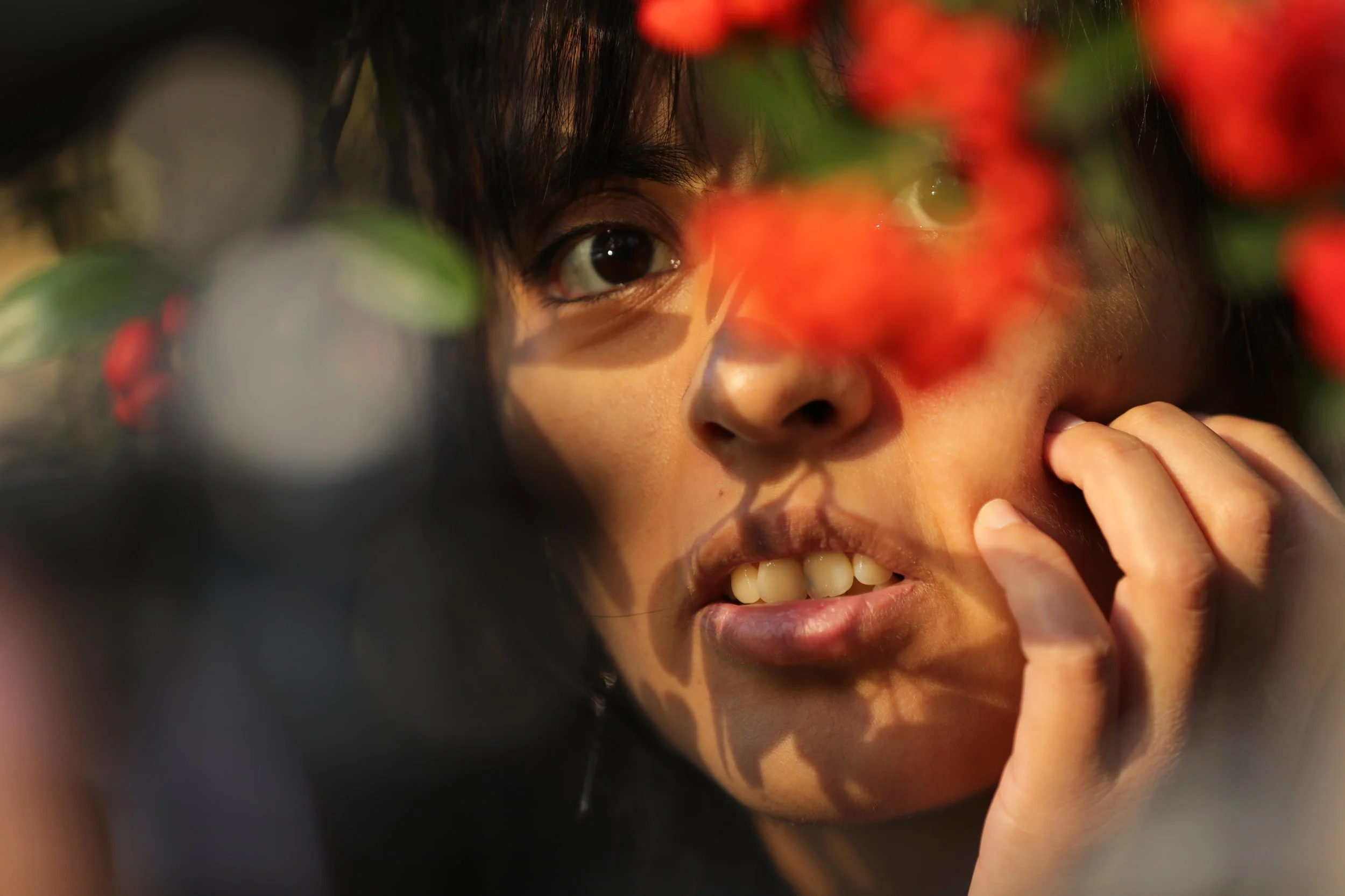 Close-up of a person with dark hair and tan skin looking through blurred red flowers, touching their face with one hand.