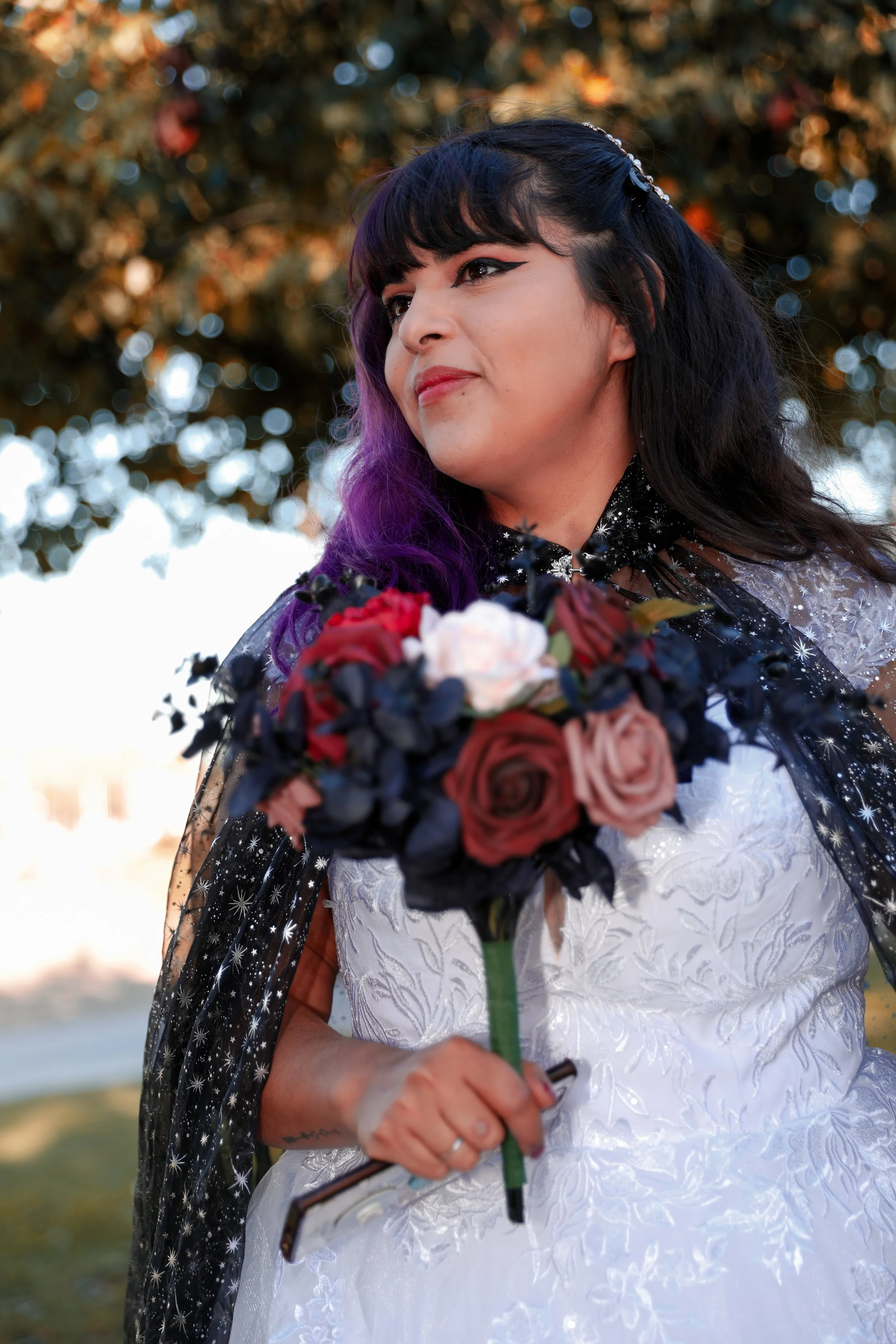 Woman with purple and black hair in a white lace dress holds a bouquet of red, pink, and white roses outdoors.