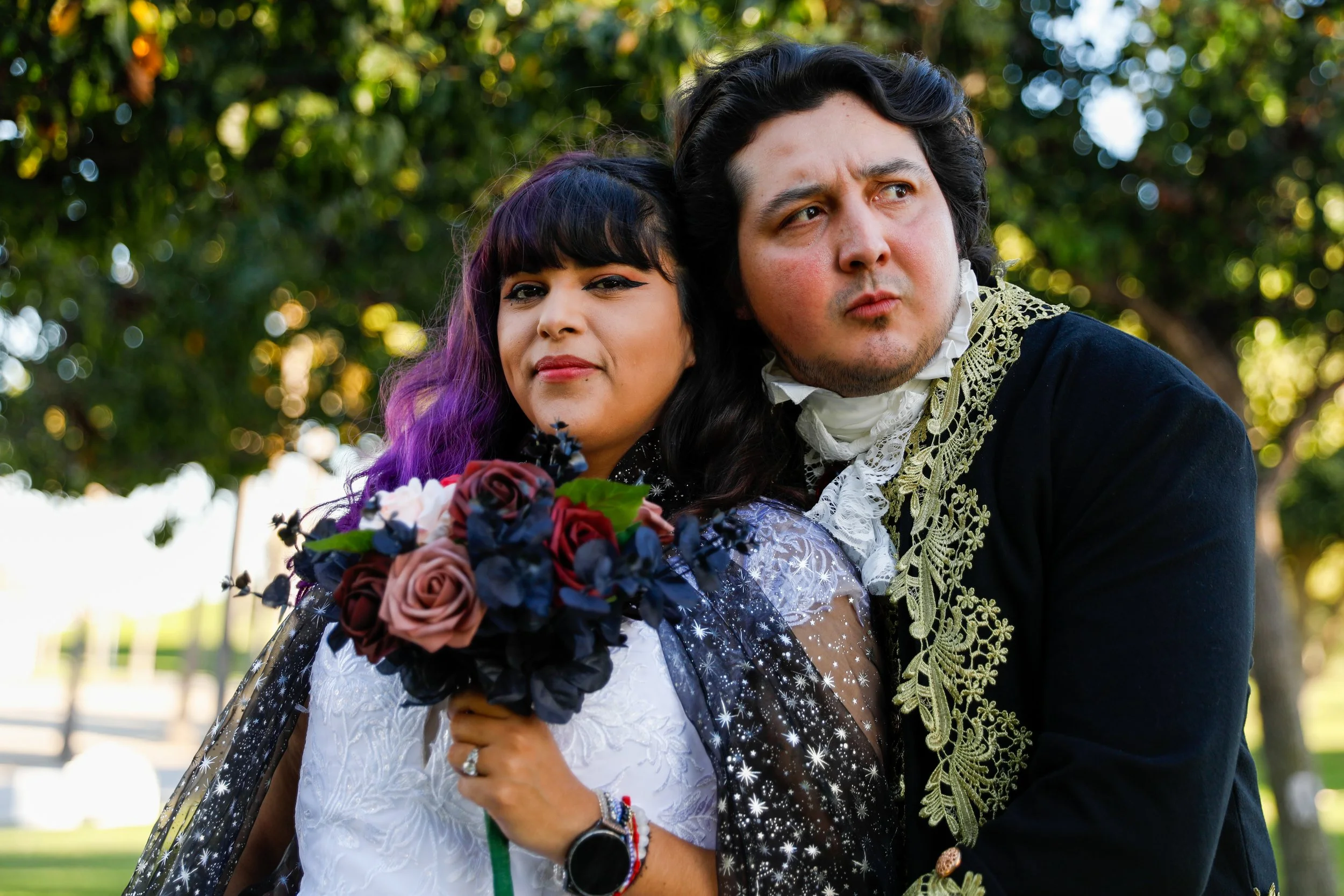 Couple dressed in renaissance-style costumes outdoors. The woman has black hair with purple streaks, and holds a bouquet of red, purple, and blue flowers. The man has dark, curly hair and is wearing a black coat with ornate gold embroidery, a white ruffled shirt, and a lace cravat.
