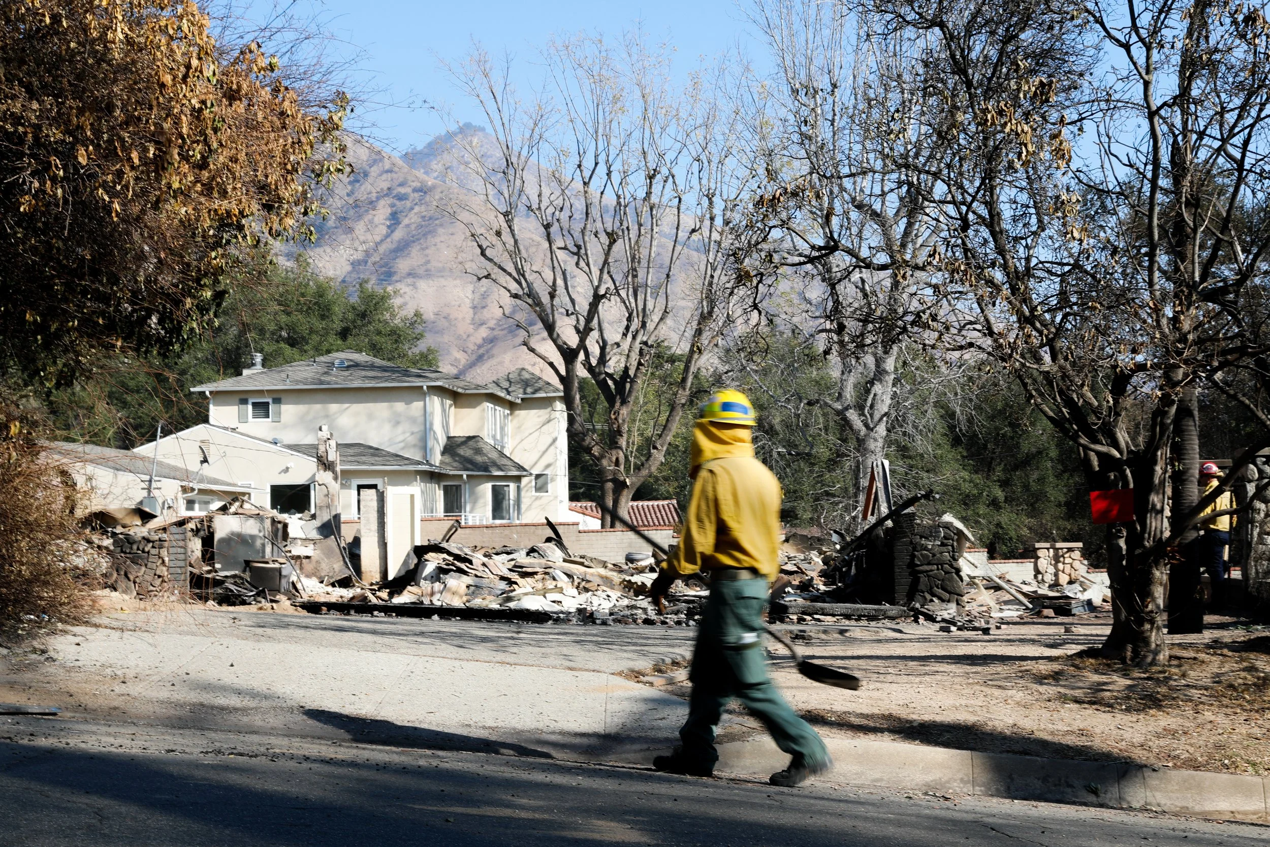 Firefighter walking past the remains of a burned house with damaged trees and mountain in the background.