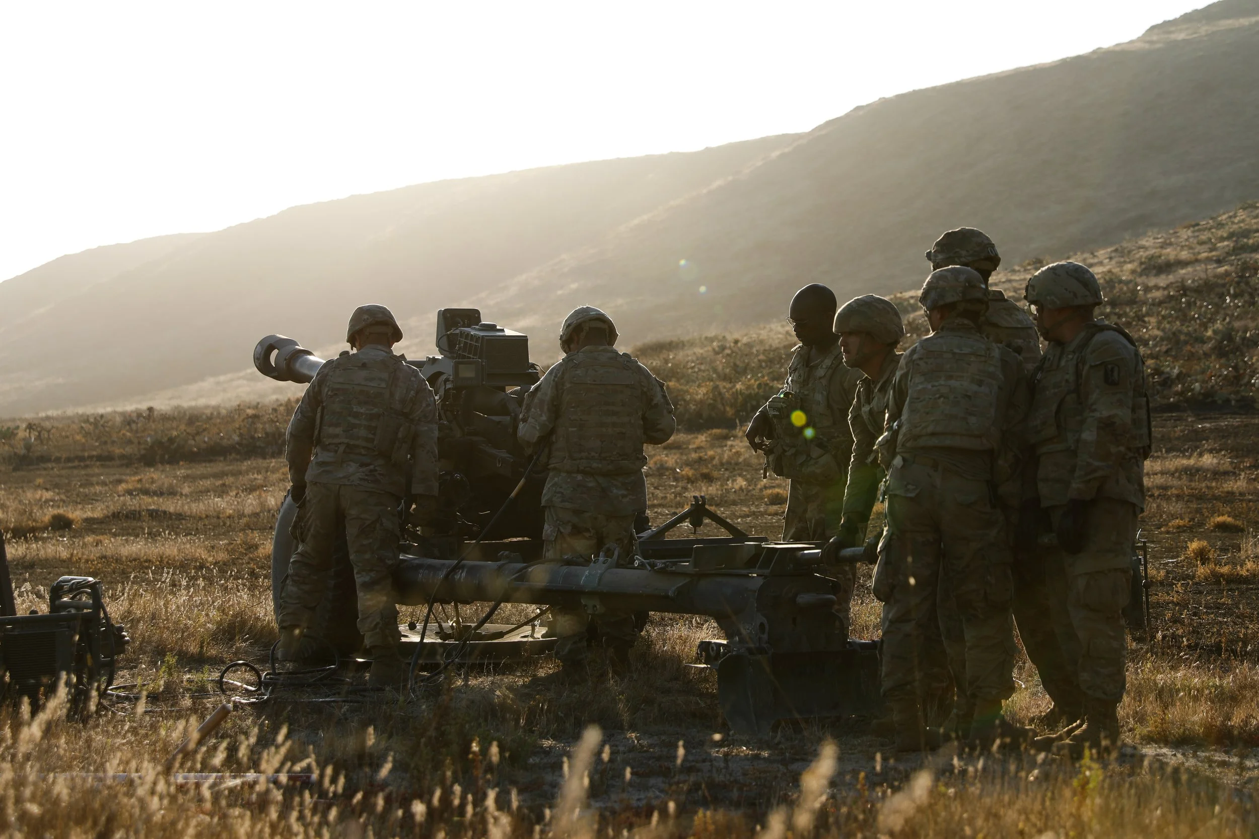 Military soldiers in camouflage uniform operating artillery during sunset in a grassy field.