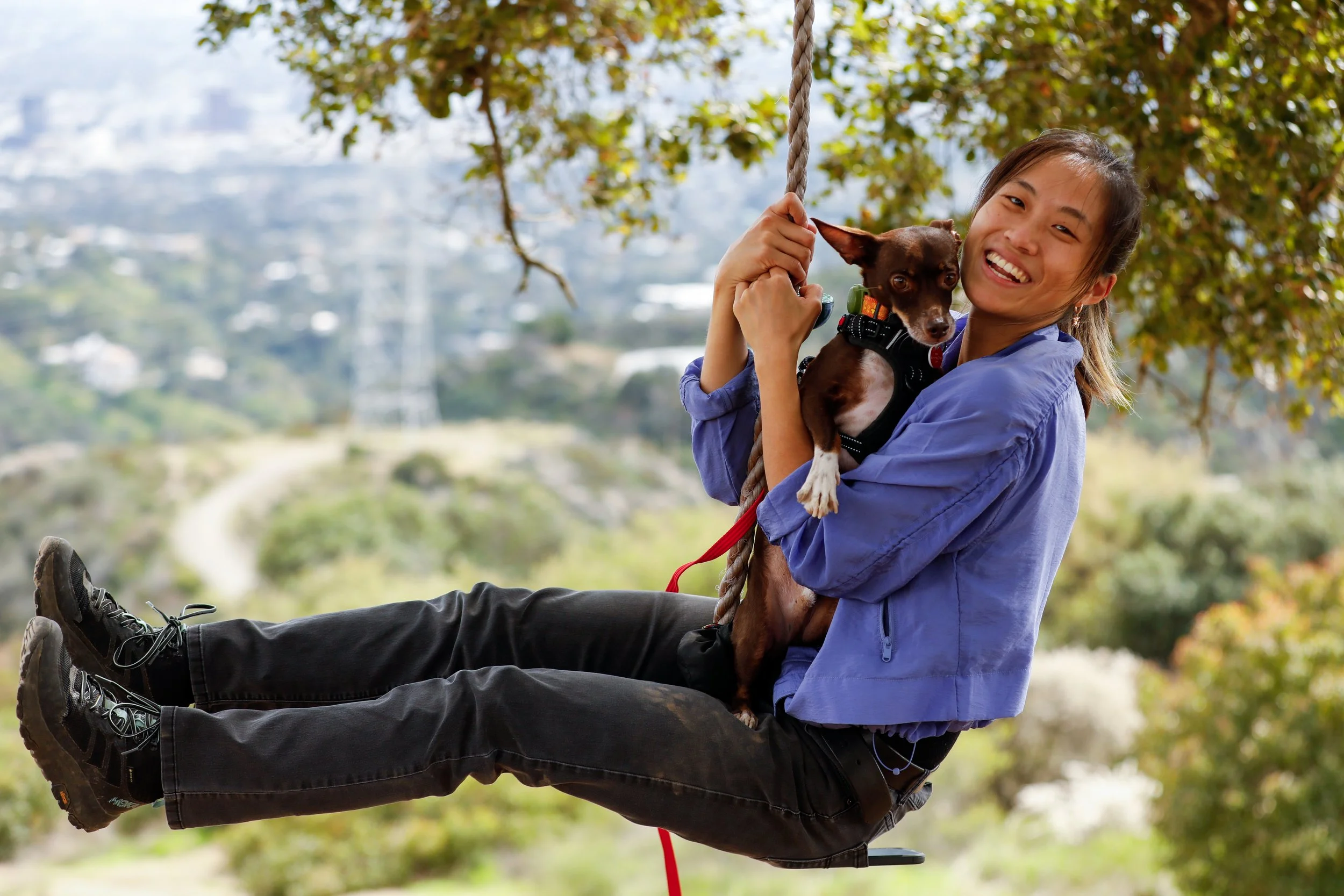 A young woman smiling and swinging on a swing with her small brown dog, outdoor setting with green trees and blurred landscape in the background.