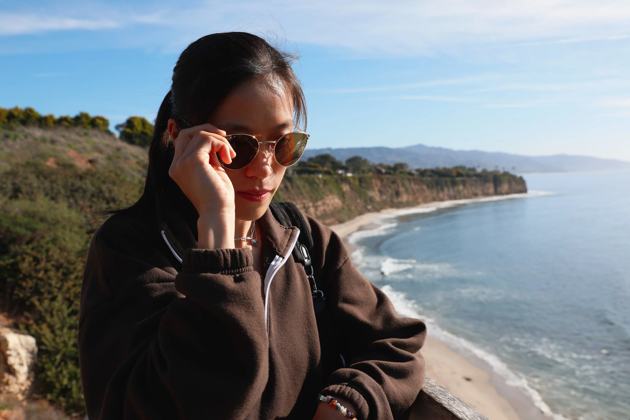 Young woman with sunglasses on a coastal overlook, with cliffs and the ocean in the background.
