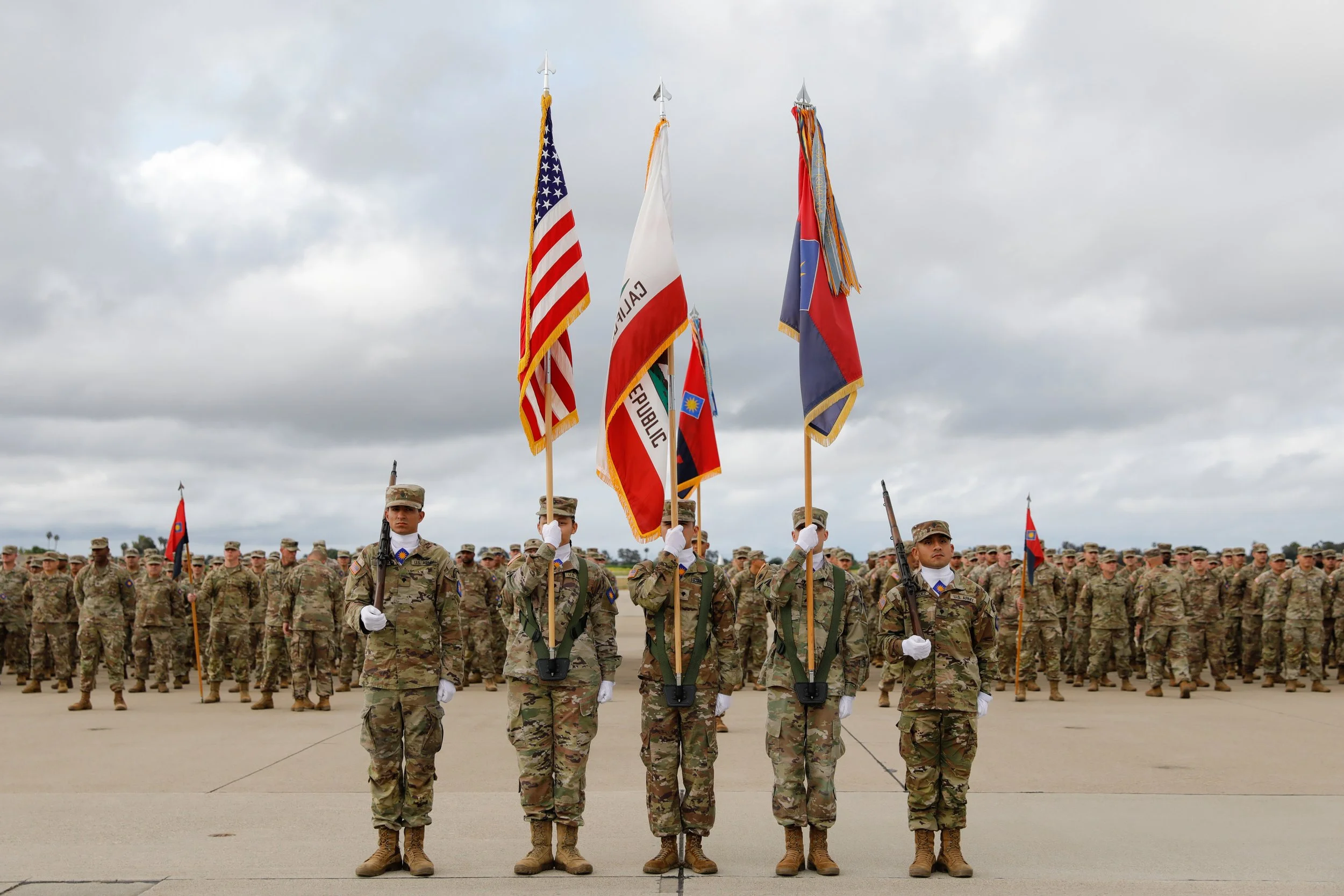 U.S. military personnel standing in formation with five soldiers in front holding flags, including the American flag, with overcast sky in the background.