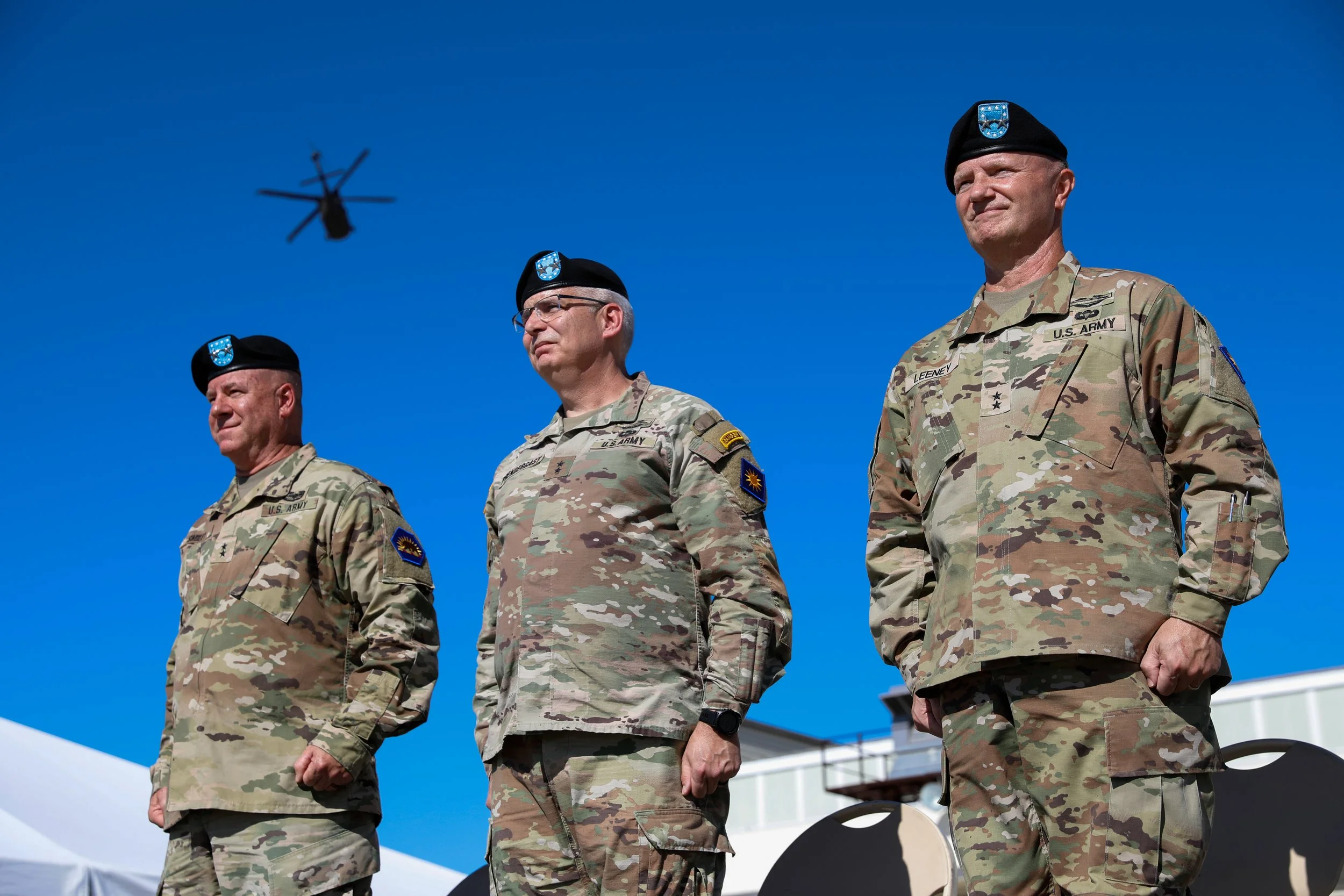 Three U.S. Army soldiers in camouflage uniforms and black berets standing outdoors during a ceremony, with a helicopter flying in the clear blue sky above them.