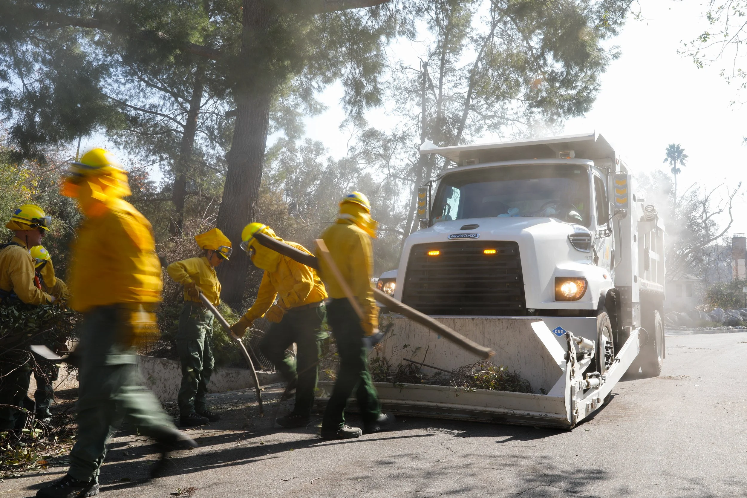 Firefighters in yellow protective clothing clearing debris from the side of a road using a large street sweeper machine, with trees in the background.