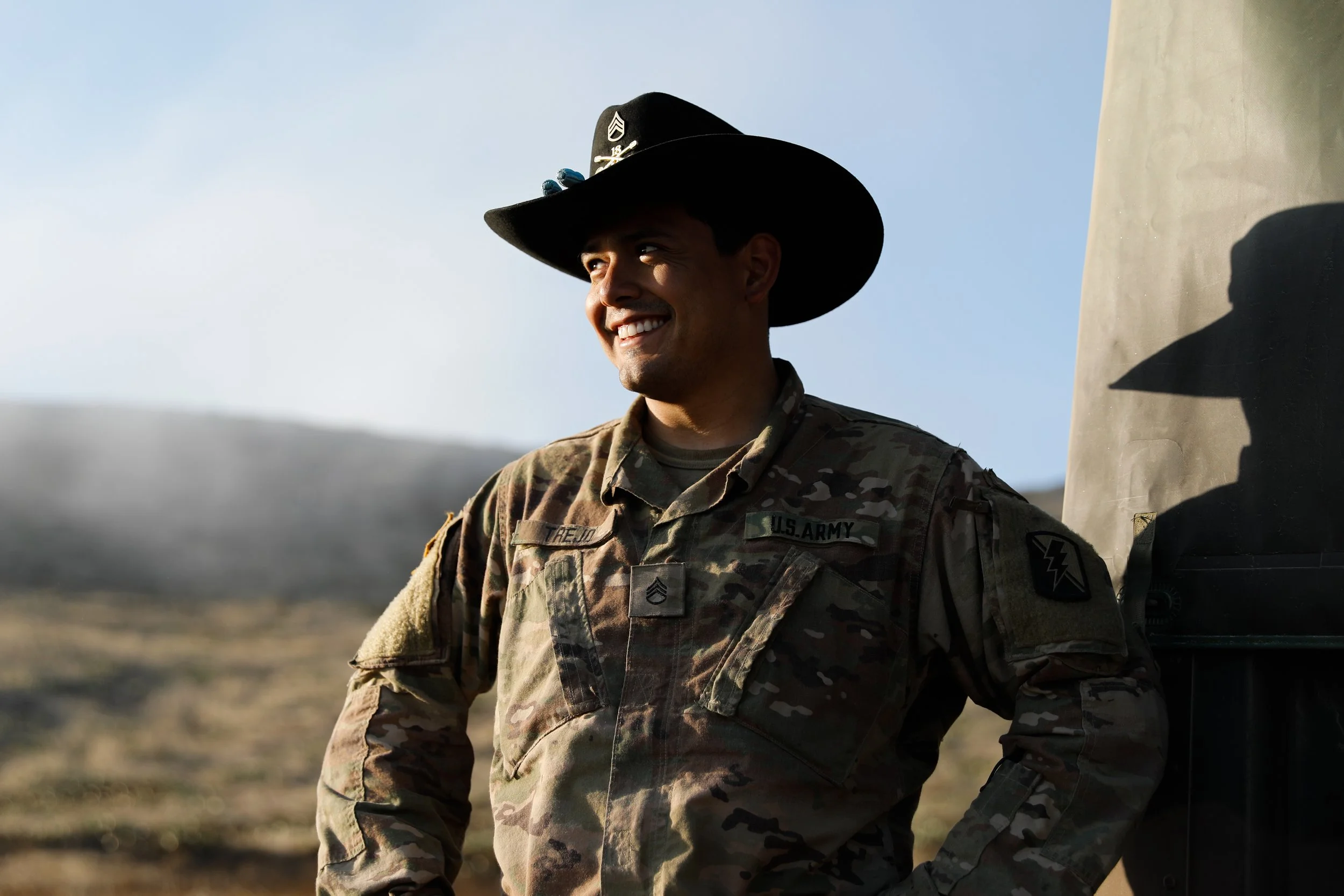 A smiling man in U.S. Army camouflage uniform and a large black cowboy hat standing outdoors with a blurred landscape and blue sky in the background.