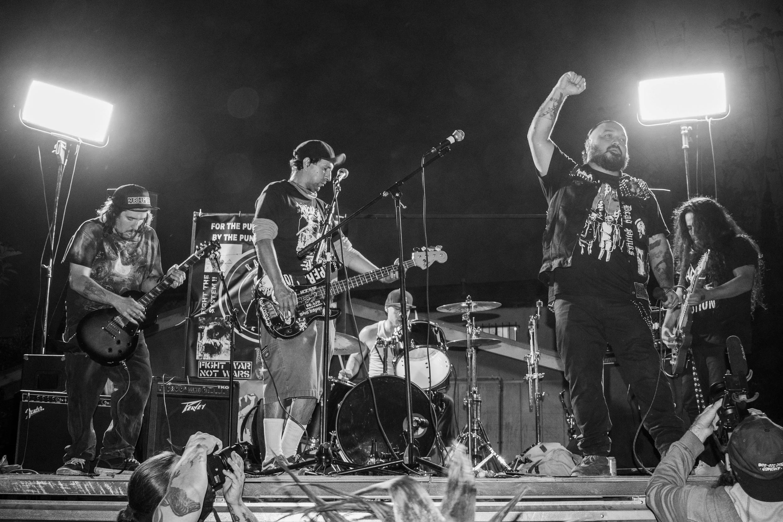 A black and white photo of a live band performing on stage, with four musicians playing guitars and drums, and a lead singer raising a fist.