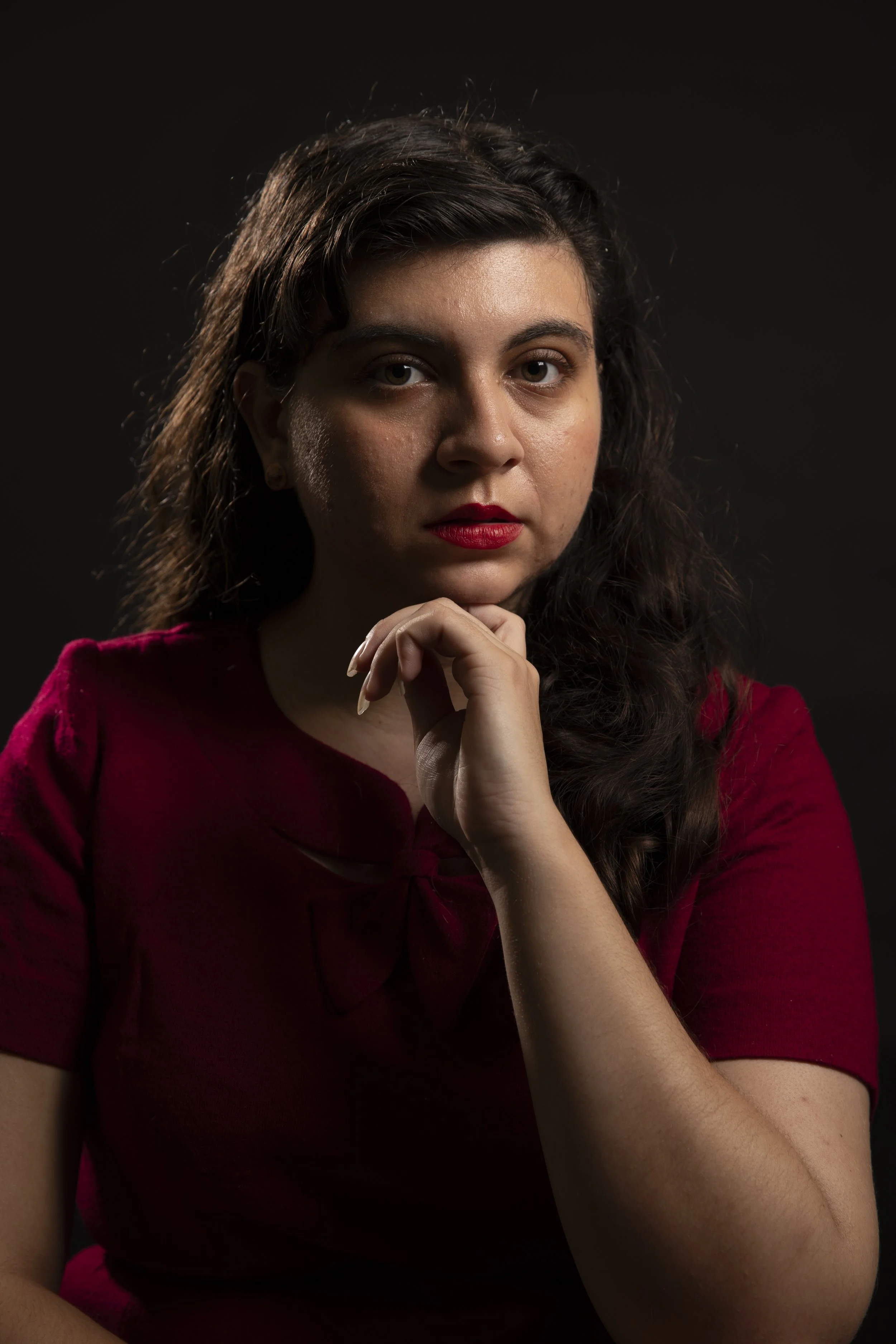 A young woman with curly dark hair, wearing a red top with a bow, poses against a black background, resting her chin on her hand and looking at the camera.