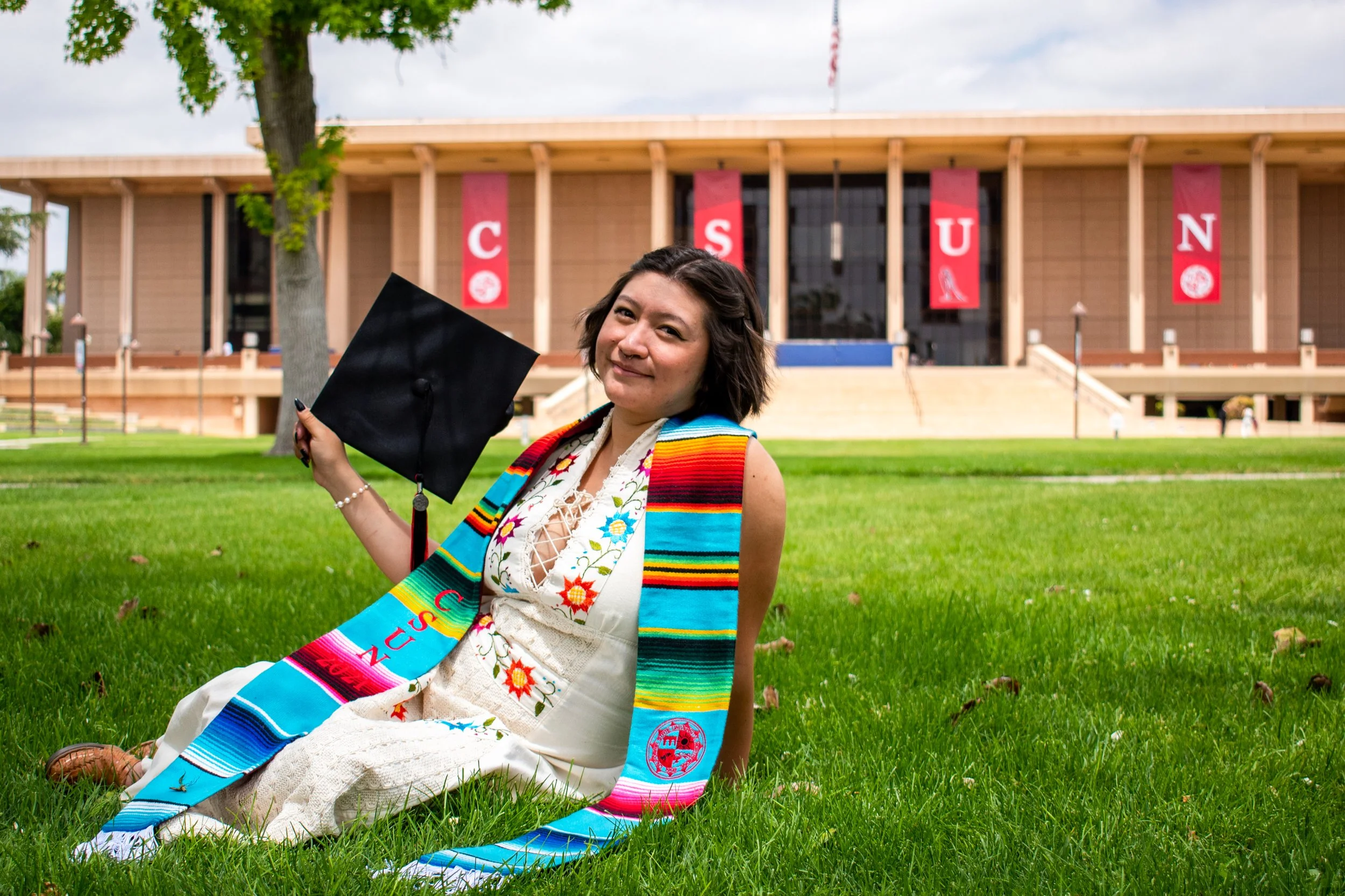 A woman sitting on the grass in graduation attire, holding a graduation cap, with a university building in the background.