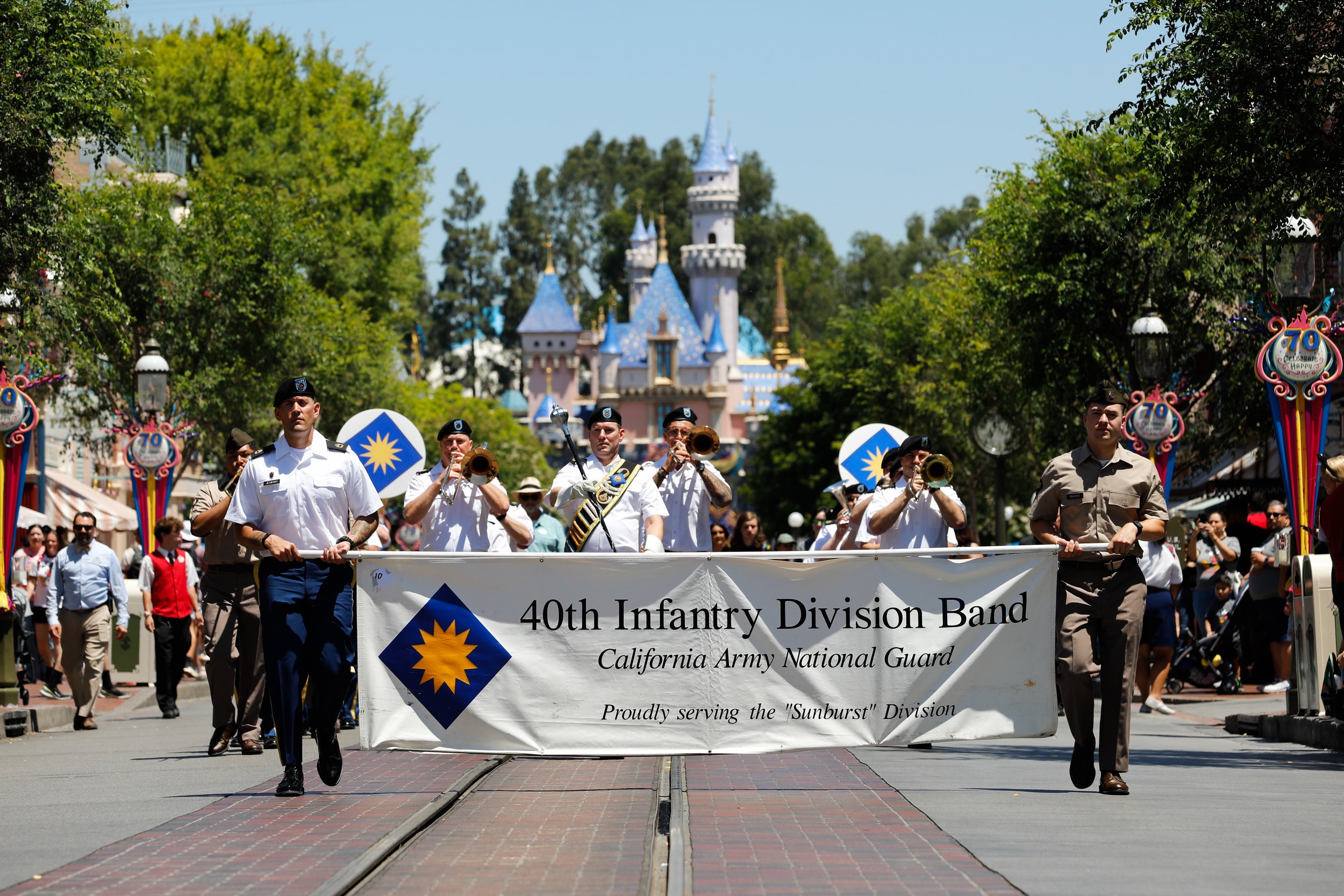 A procession of military band members marching in a parade on Main Street with the Sleeping Beauty Castle in the background. The band is from the 40th Infantry Division Band, California Army National Guard, serving the Sunburst Division. The parade features flags and spectators watching on both sides of the street.