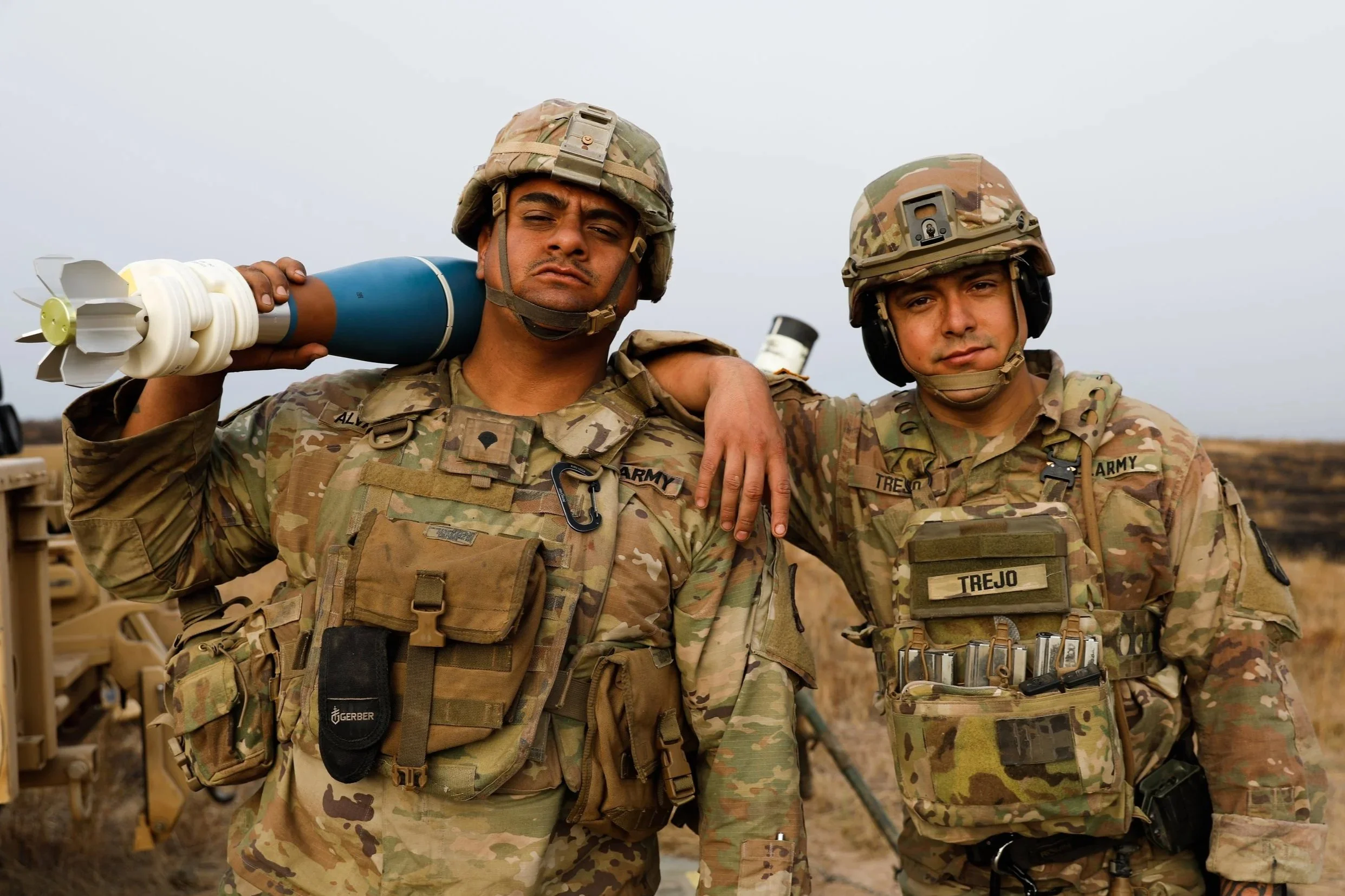 Two soldiers in camouflage uniforms and helmets standing close together outdoors, with one carrying a mortar round over his shoulder. They have serious expressions.