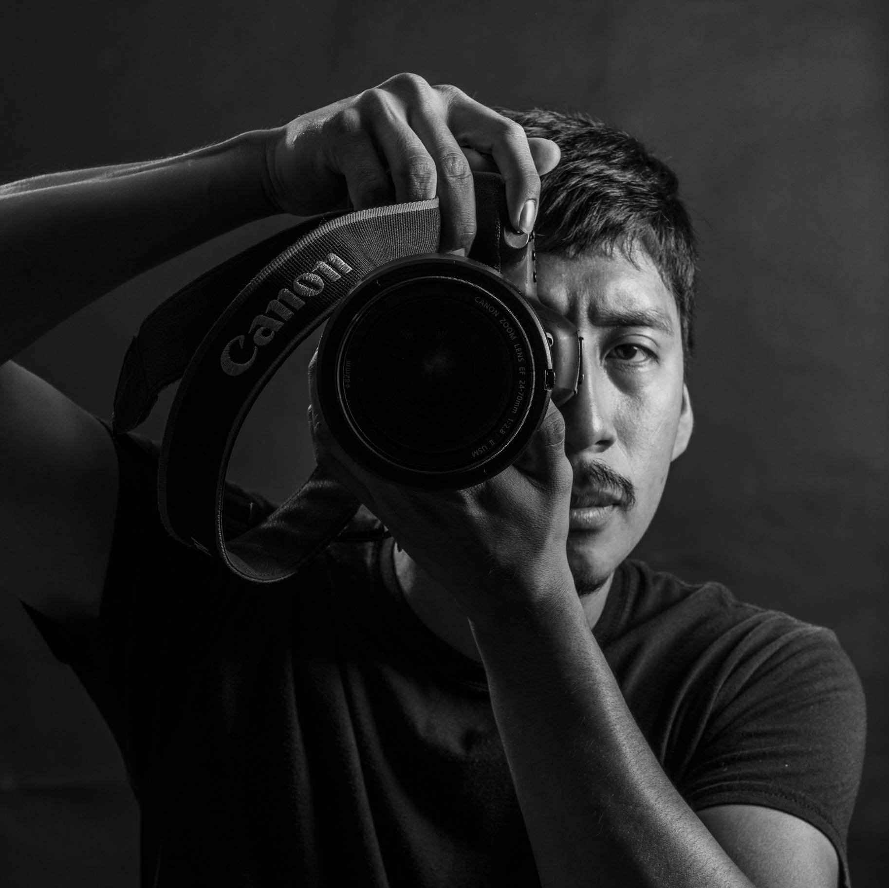 A young man takes a self-portrait in black and white, holding a Canon camera up to his eye with one hand and supporting the lens with the other, against a dark background.