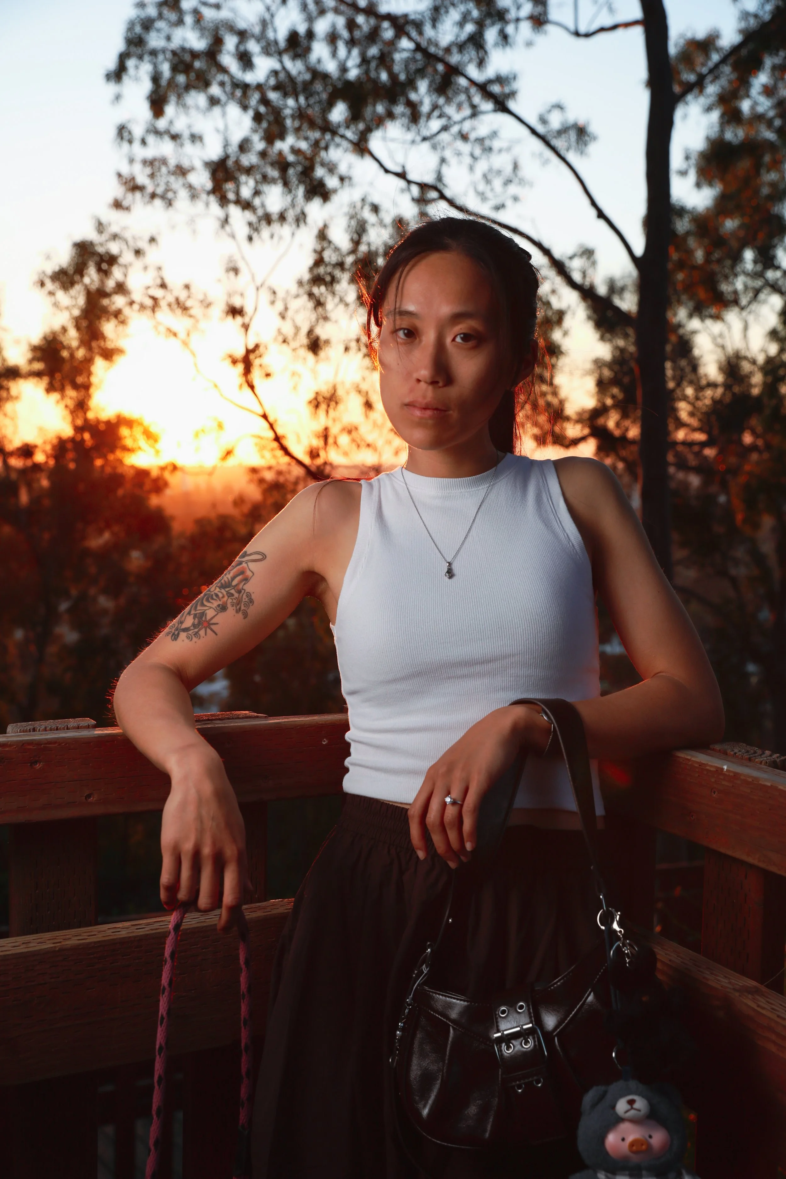 A woman standing outdoors on a wooden platform at sunset, wearing a white sleeveless top, black skirt, and holding a black handbag and a pink rope leash, with trees in the background.