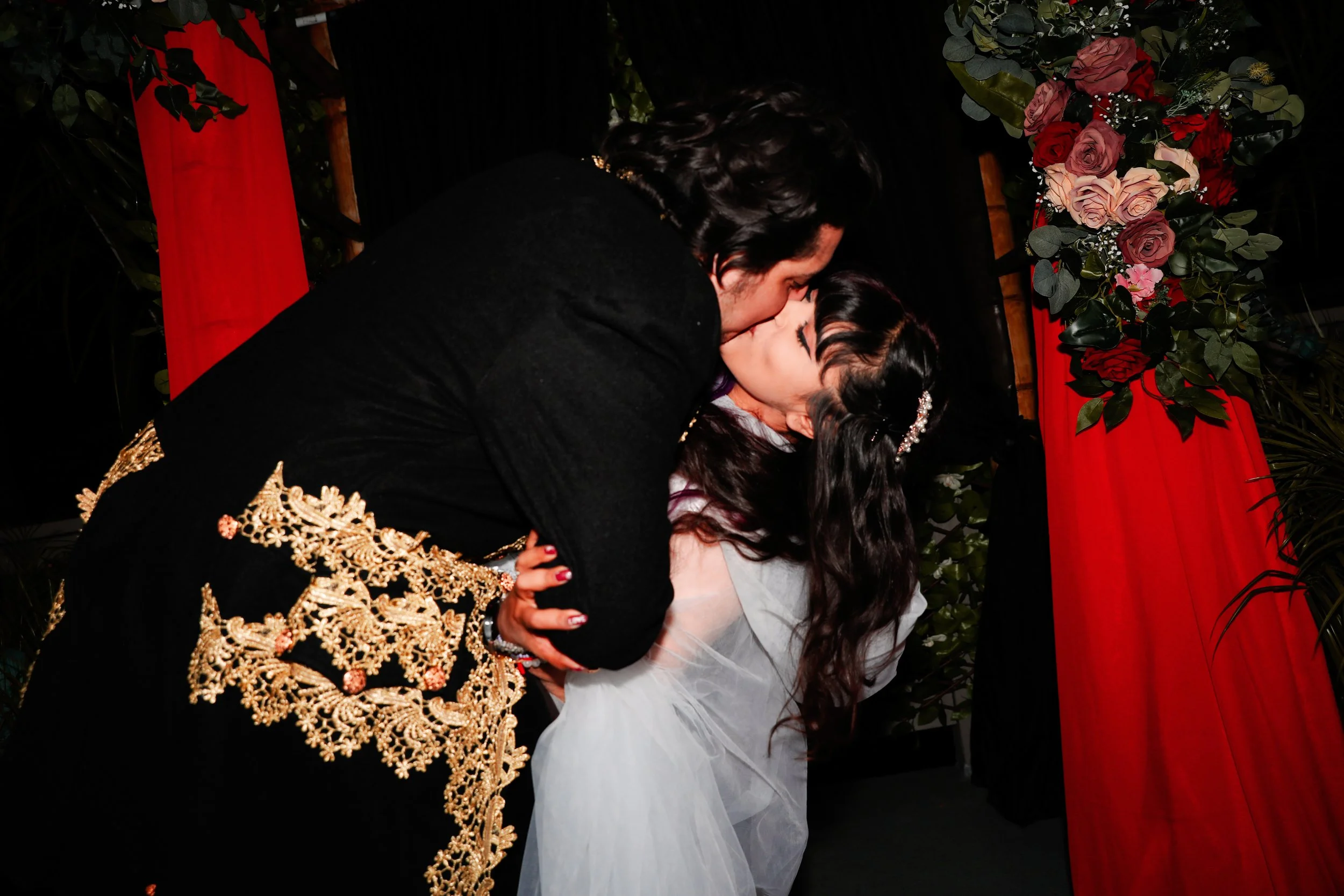 A man and a girl are sharing a kiss at a celebratory event, with floral decorations and red and black drapes in the background.