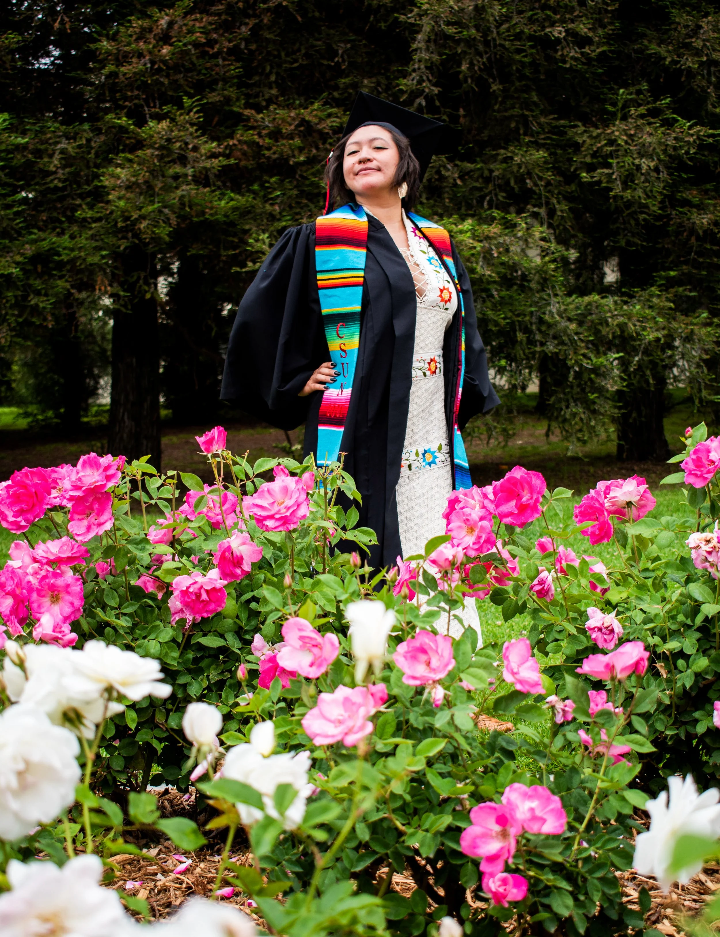 A woman in graduation cap and gown standing in a garden of pink and white flowers, with tall trees in the background.