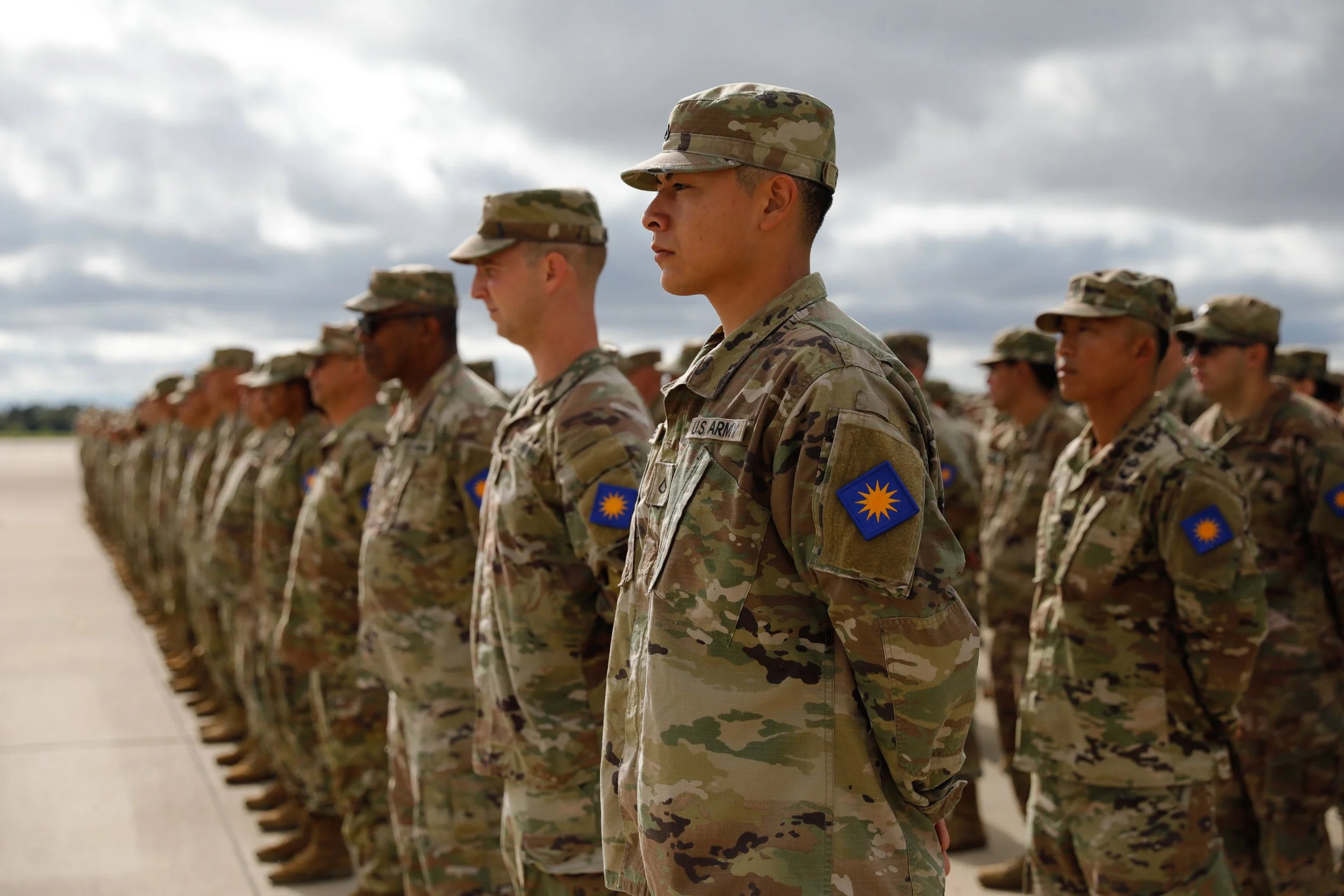 Group of soldiers in camouflage uniforms standing in formation outdoor under cloudy sky.