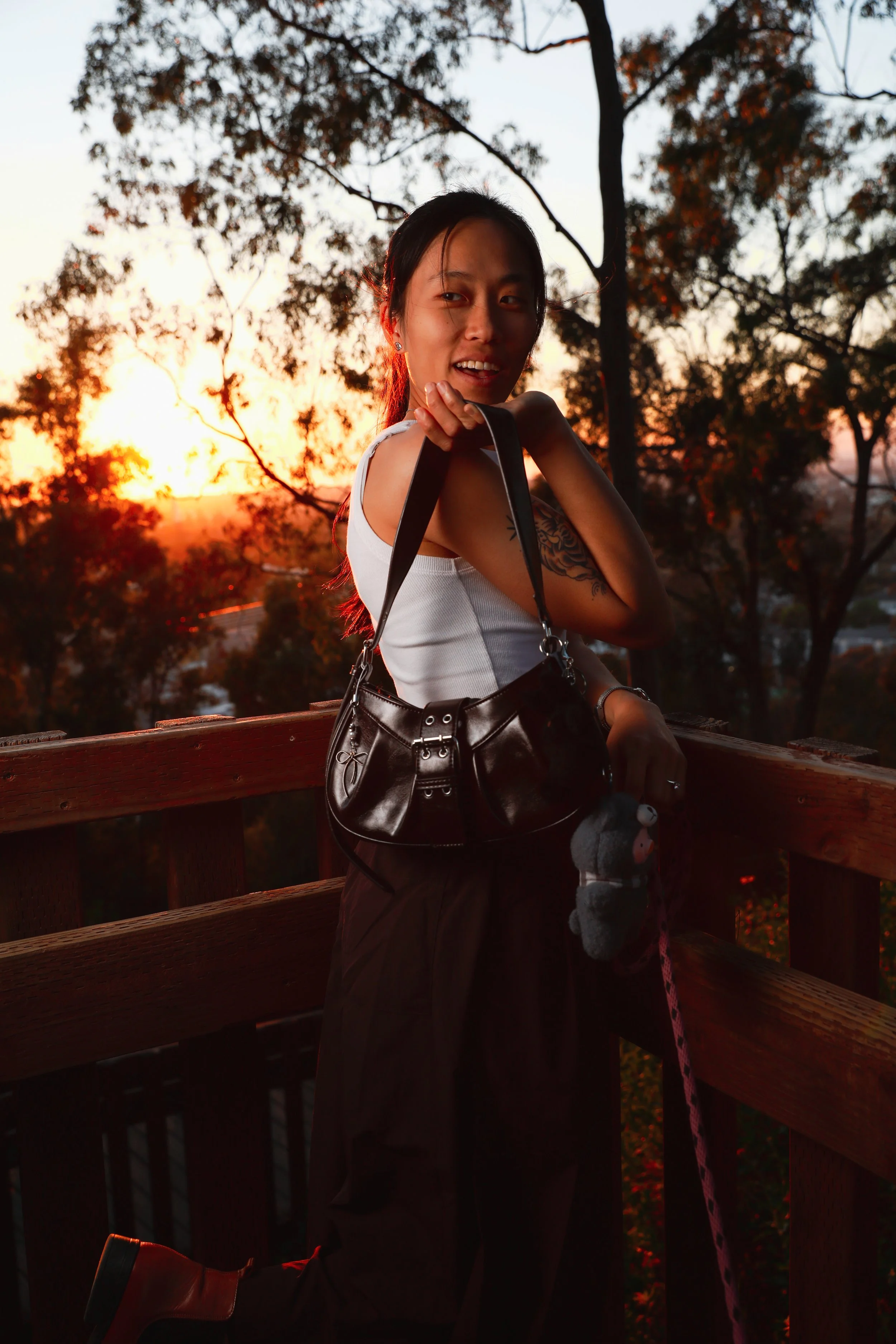 A woman holding a black handbag and a small stuffed toy, standing on a wooden deck during sunset with trees in the background.