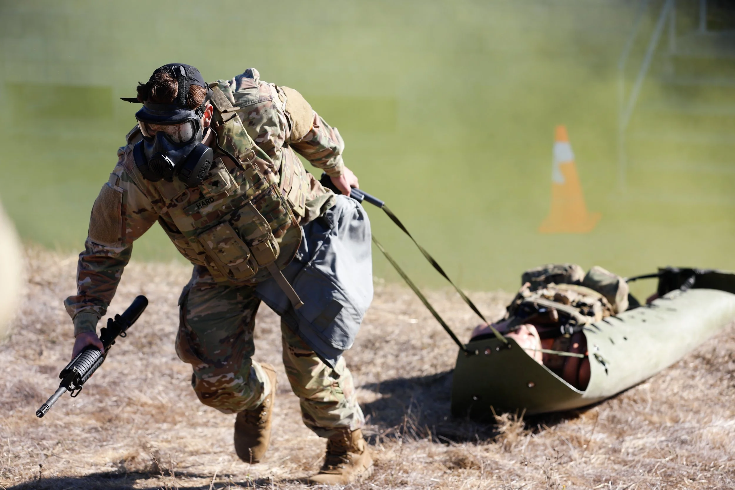 A person in camouflage military clothing, wearing a mask, goggles, and a cap, holding a rifle, pulling a kayak with gear on a grassy shoreline near water with an orange cone in the background.