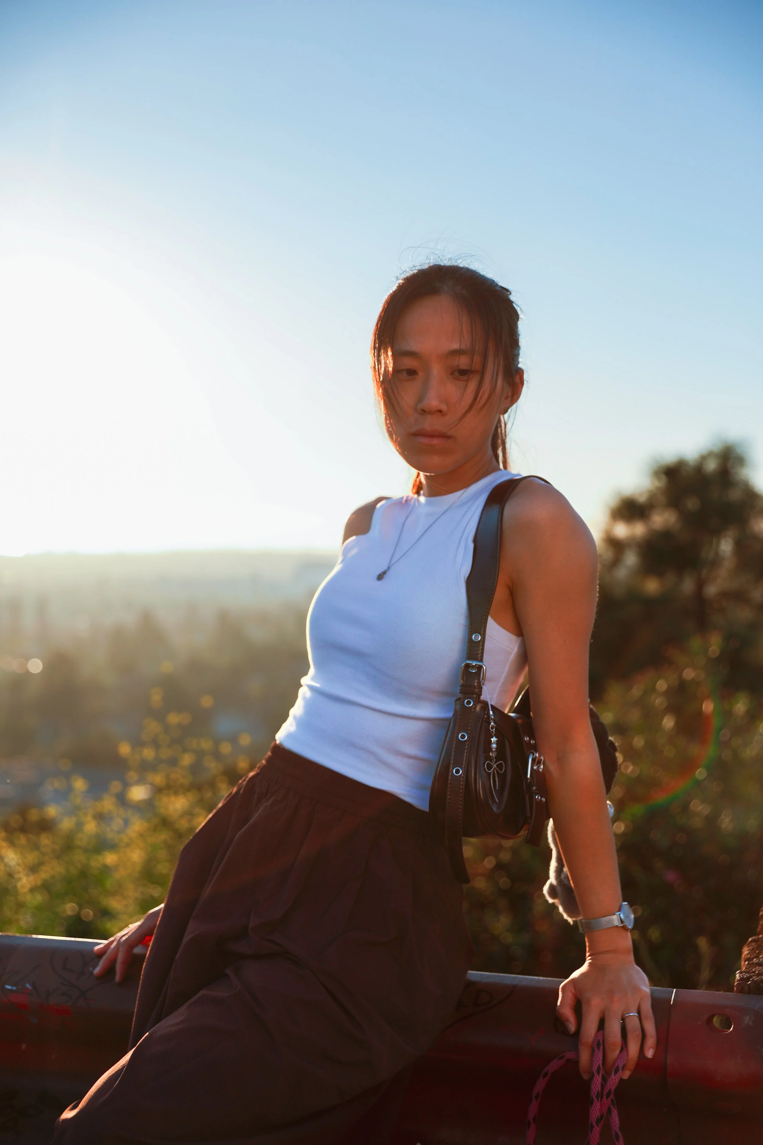 A young woman sitting on a barrier outdoors during sunset, wearing a white tank top, black pants, a watch, and carrying a small black bag.