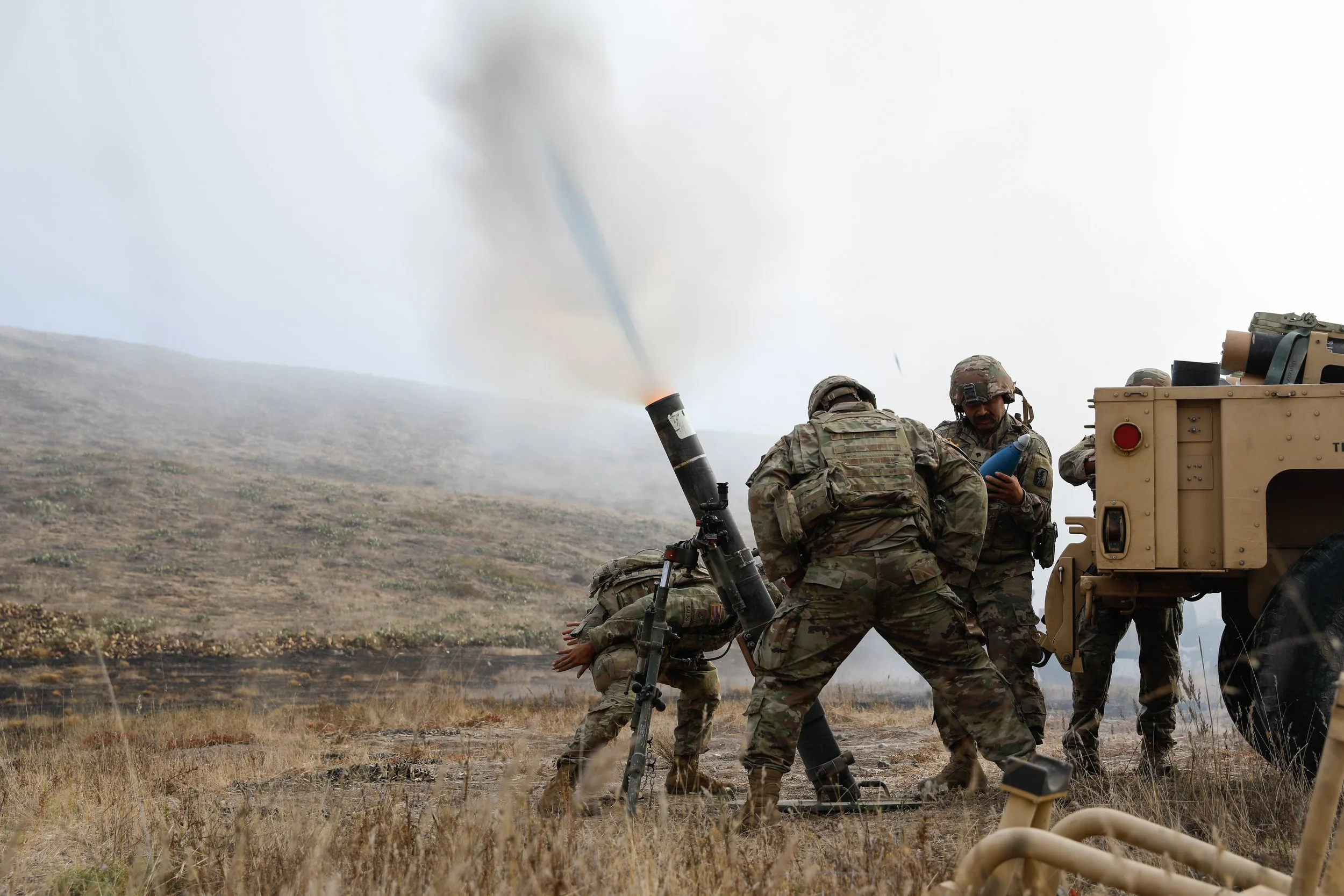 Military personnel operating a mortar, with smoke and a field in the background.