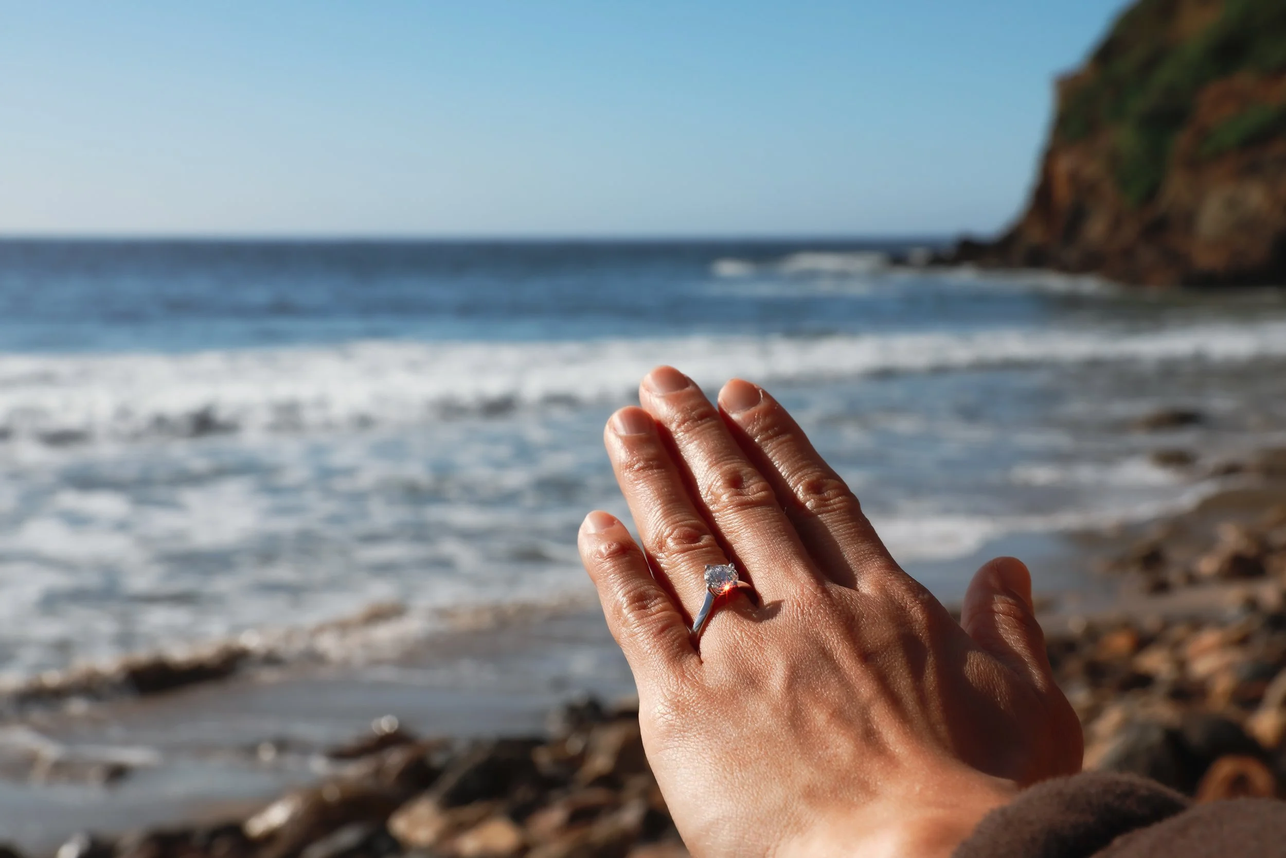 Close-up of a hand with an engagement ring, held up against a beach with waves and a rocky cliff in the background.