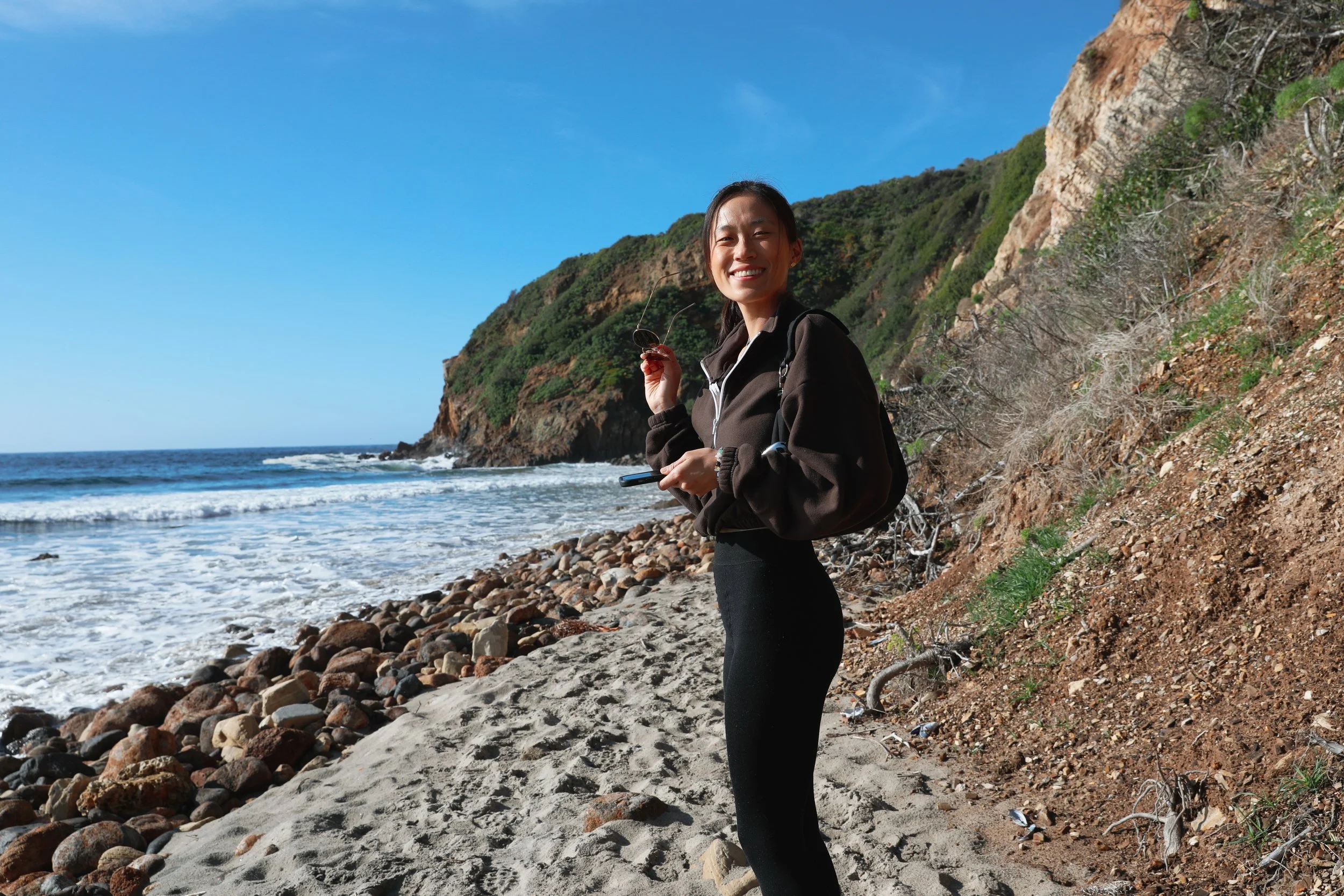 A woman smiling on a rocky beach with a cliffside and ocean waves in the background.