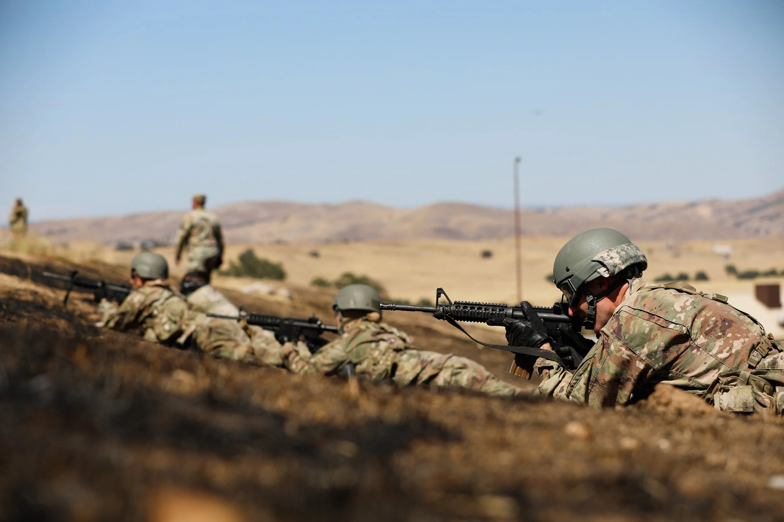 Soldiers in camouflage uniforms and helmets lying prone with rifles in a dry, hilly landscape during daytime.