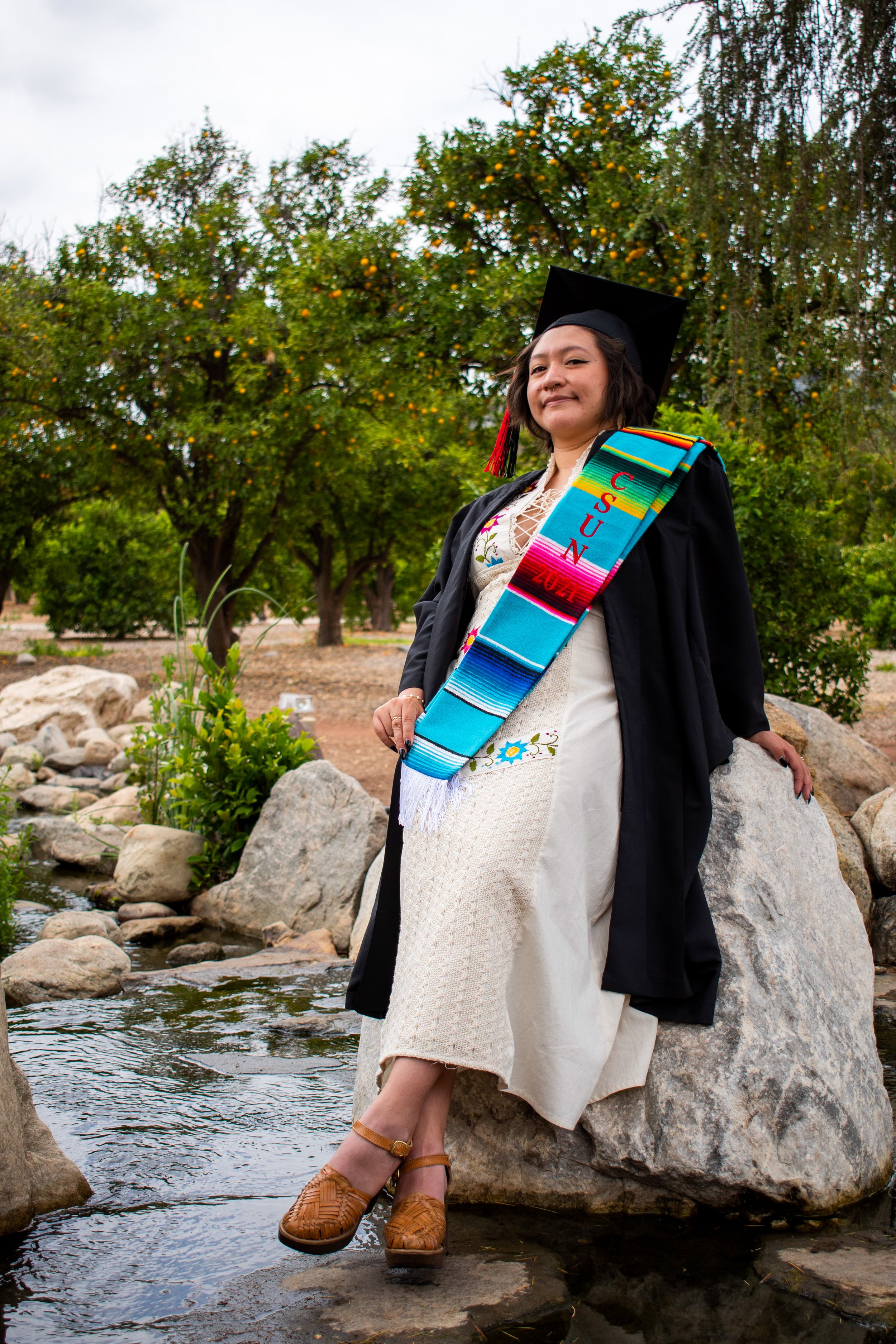 A woman in a graduation cap and gown sitting on a large rock in a natural setting with trees and a small stream.