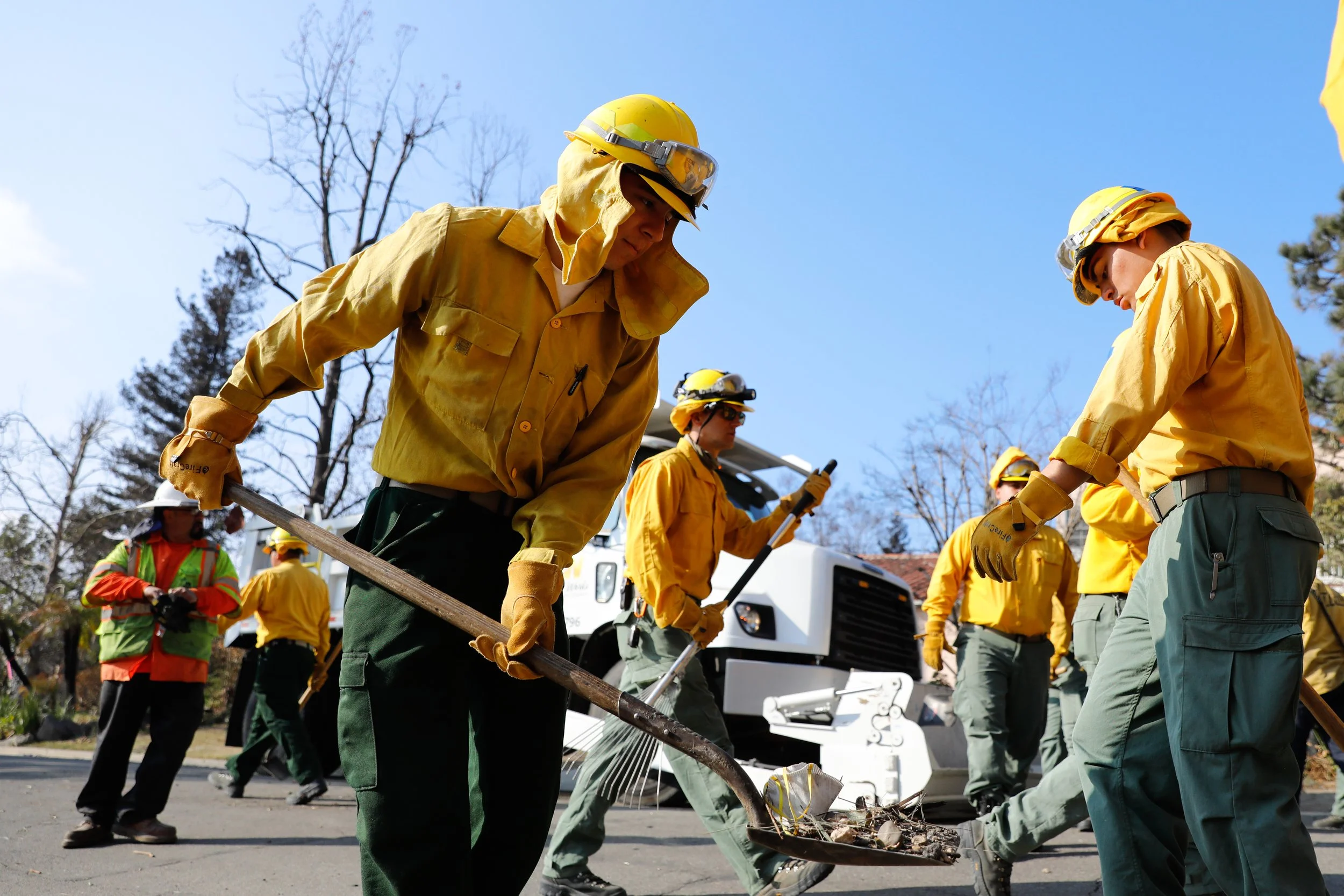 Firefighters in yellow uniforms and helmets working on debris cleanup on a sunny day.