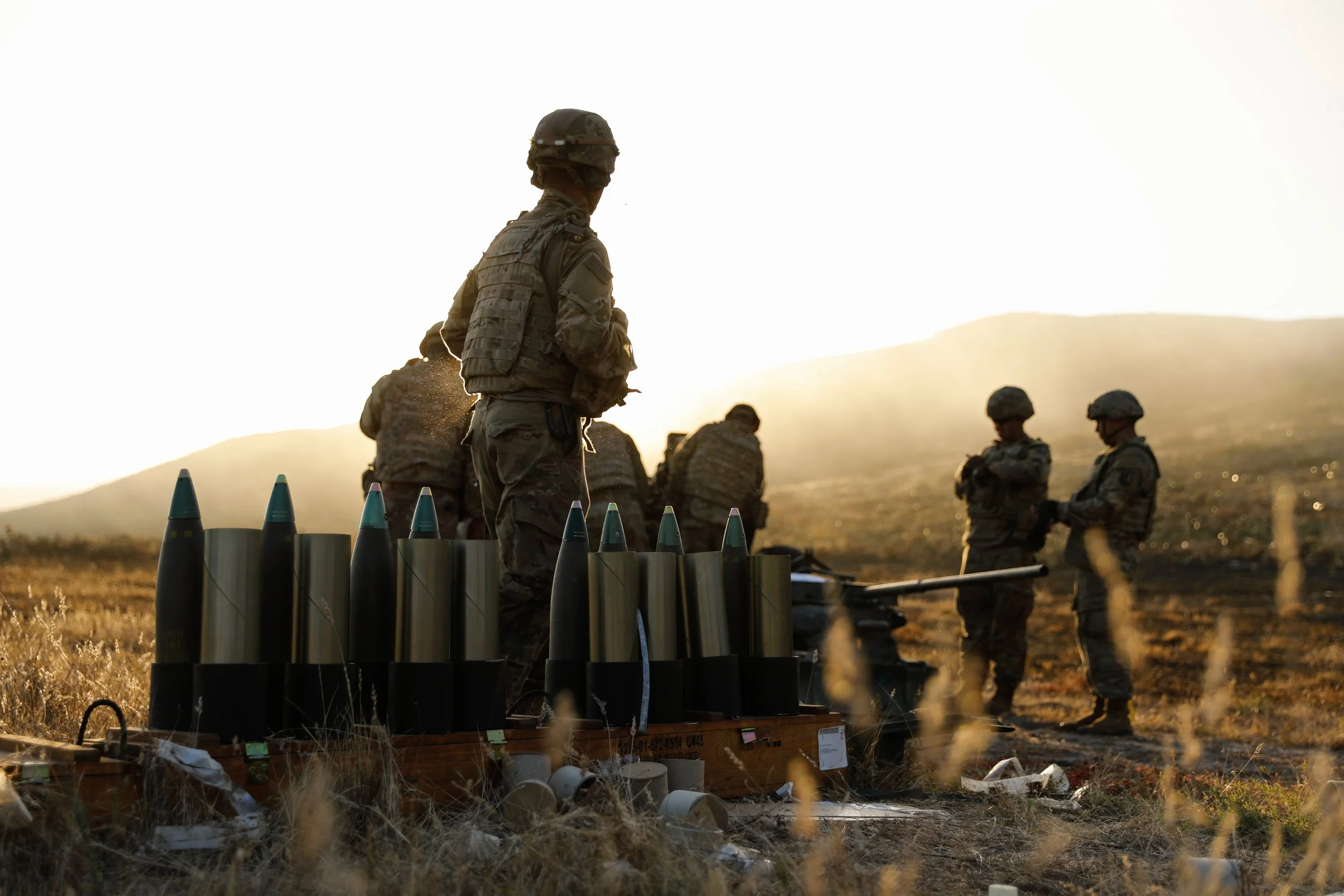 Military personnel setting up or inspecting artillery shells and equipment outdoors during sunset.