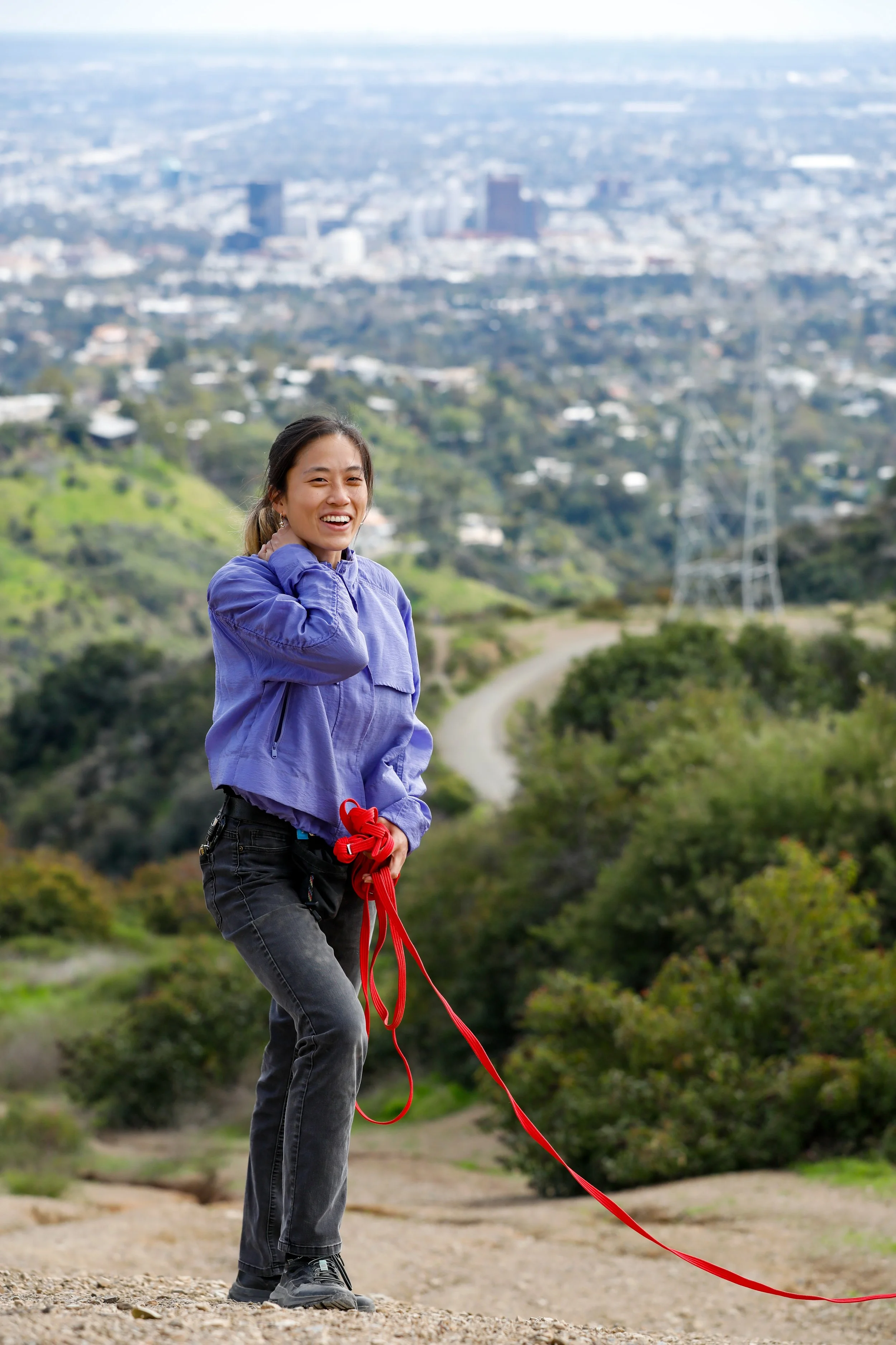 A woman in a purple jacket holding a red leash, smiling on a hilly trail with greenery and cityscape in the background.
