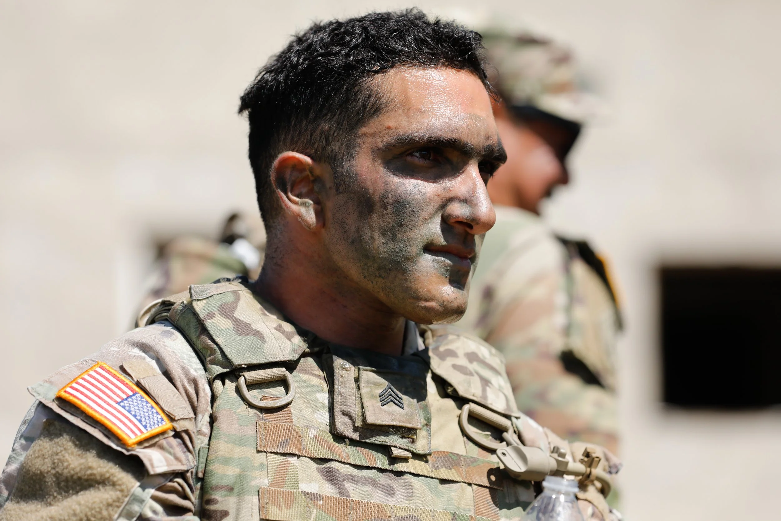 A soldier in camouflage uniform standing outdoors, with another soldier blurred in the background.